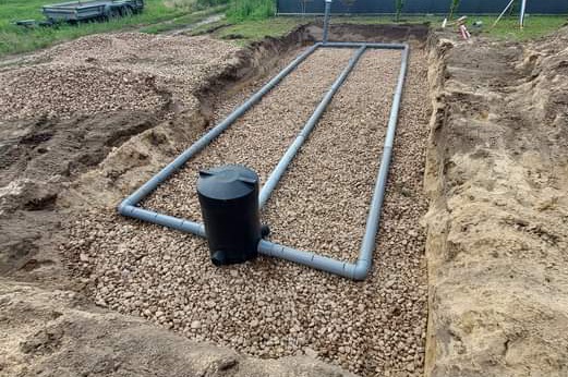 Rozłożony system rur drenarskich na żwirowym podłożu, czarna studzienka kontrolna, widok z góry na wykop z ziemią po bokach.