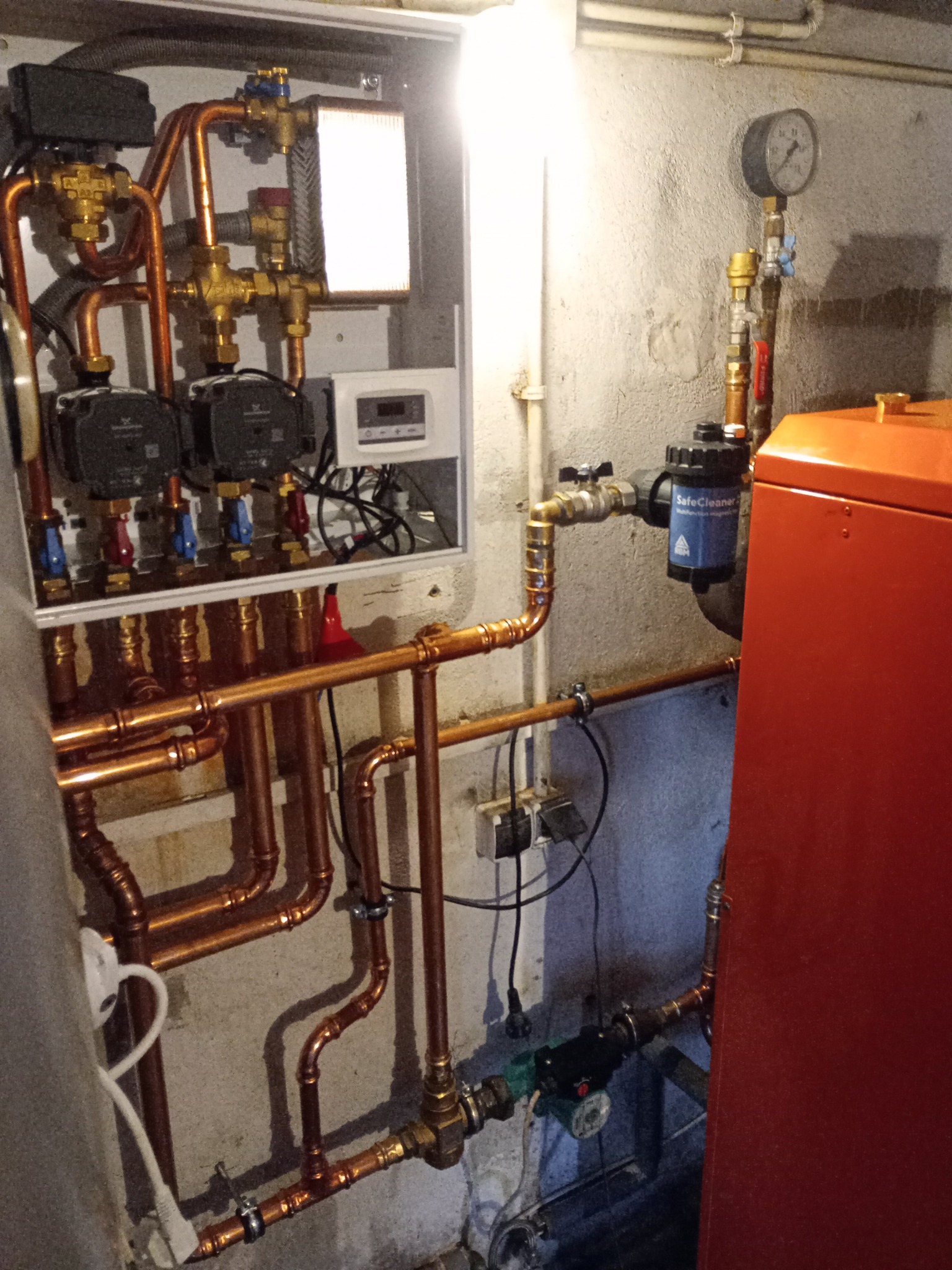 Złożona instalacja hydrauliczna z miedzianymi rurami, zaworami, pompami obiegowymi i filtrem SafeCleaner. Manometr i sterownik w tle. Widoczne elementy kotła grzewczego.