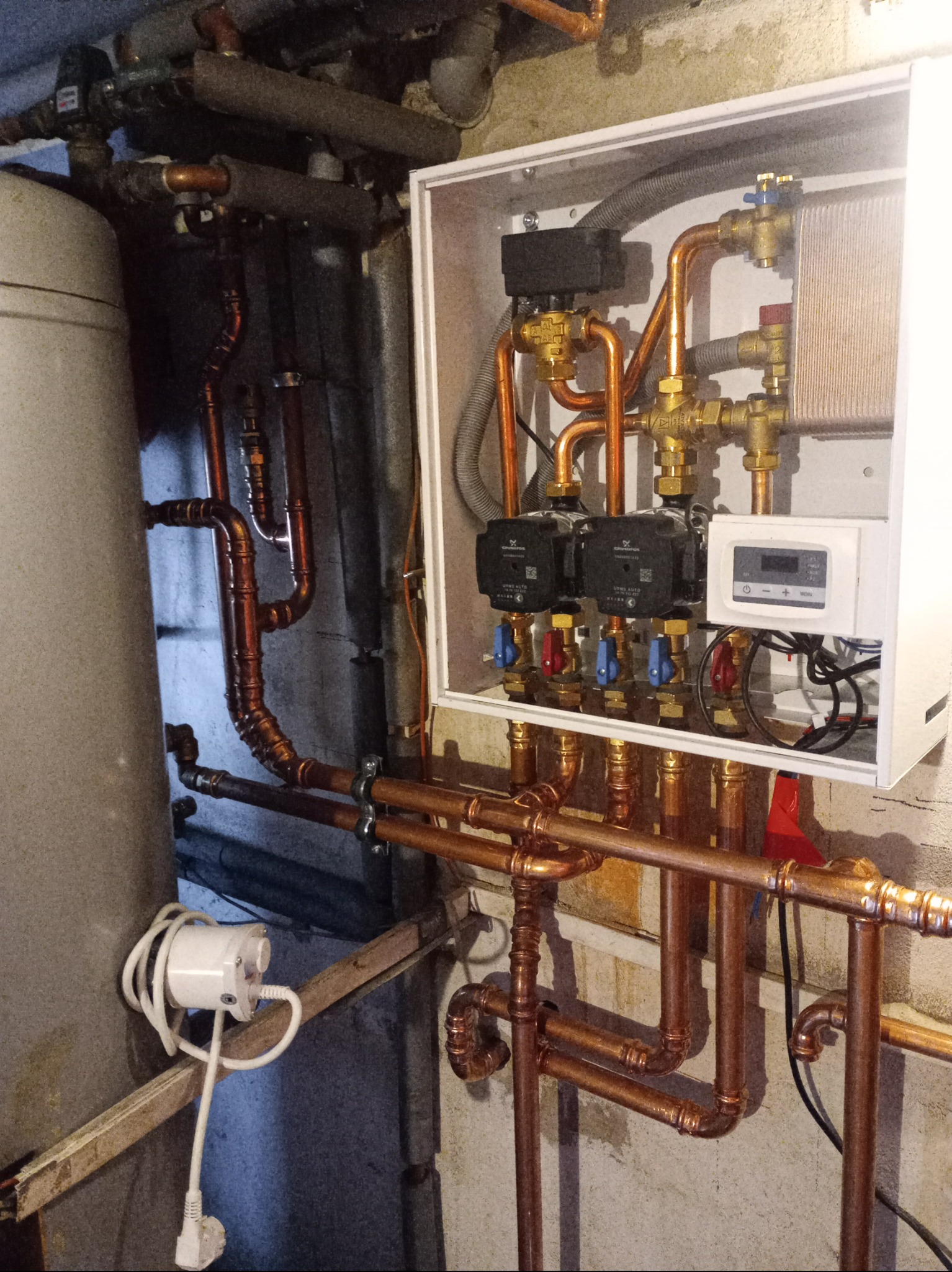 Kompleksowa instalacja grzewcza z miedzianymi rurami, pompami obiegowymi i sterownikiem w białej obudowie. Widoczny zbiornik i fragment ściany z fakturą.