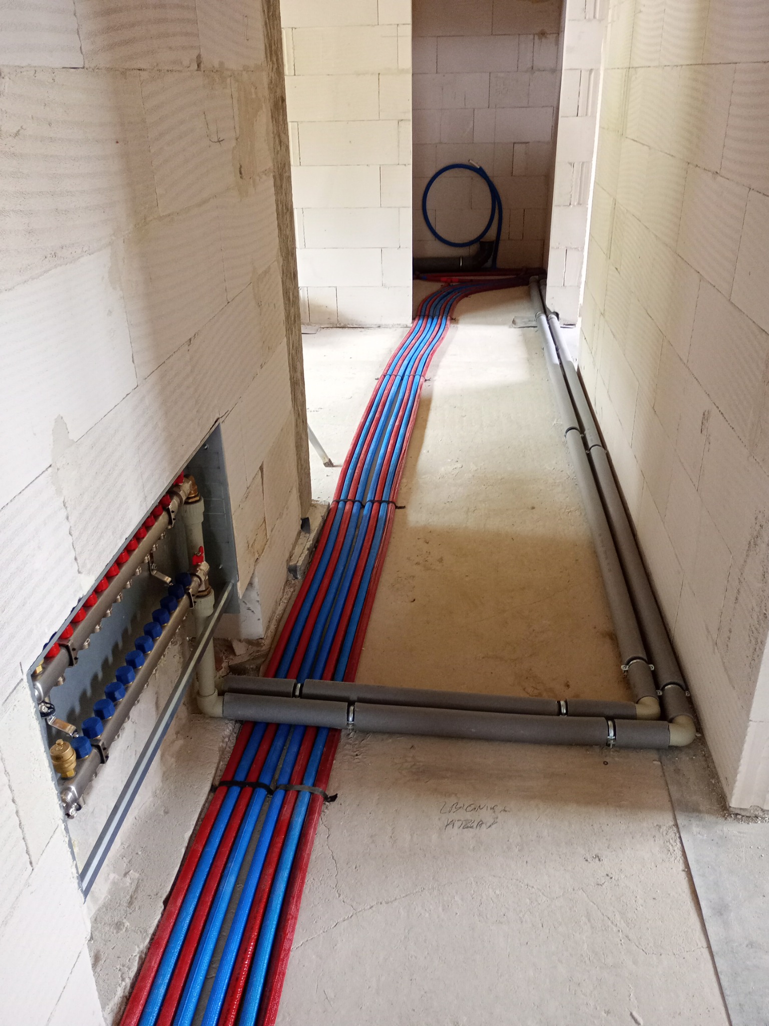 Rozdzielacz ogrzewania podłogowego z czerwono-niebieskimi rurami w surowym wnętrzu z bloczków, instalacja w trakcie realizacji, widoczne rury szare w izolacji.