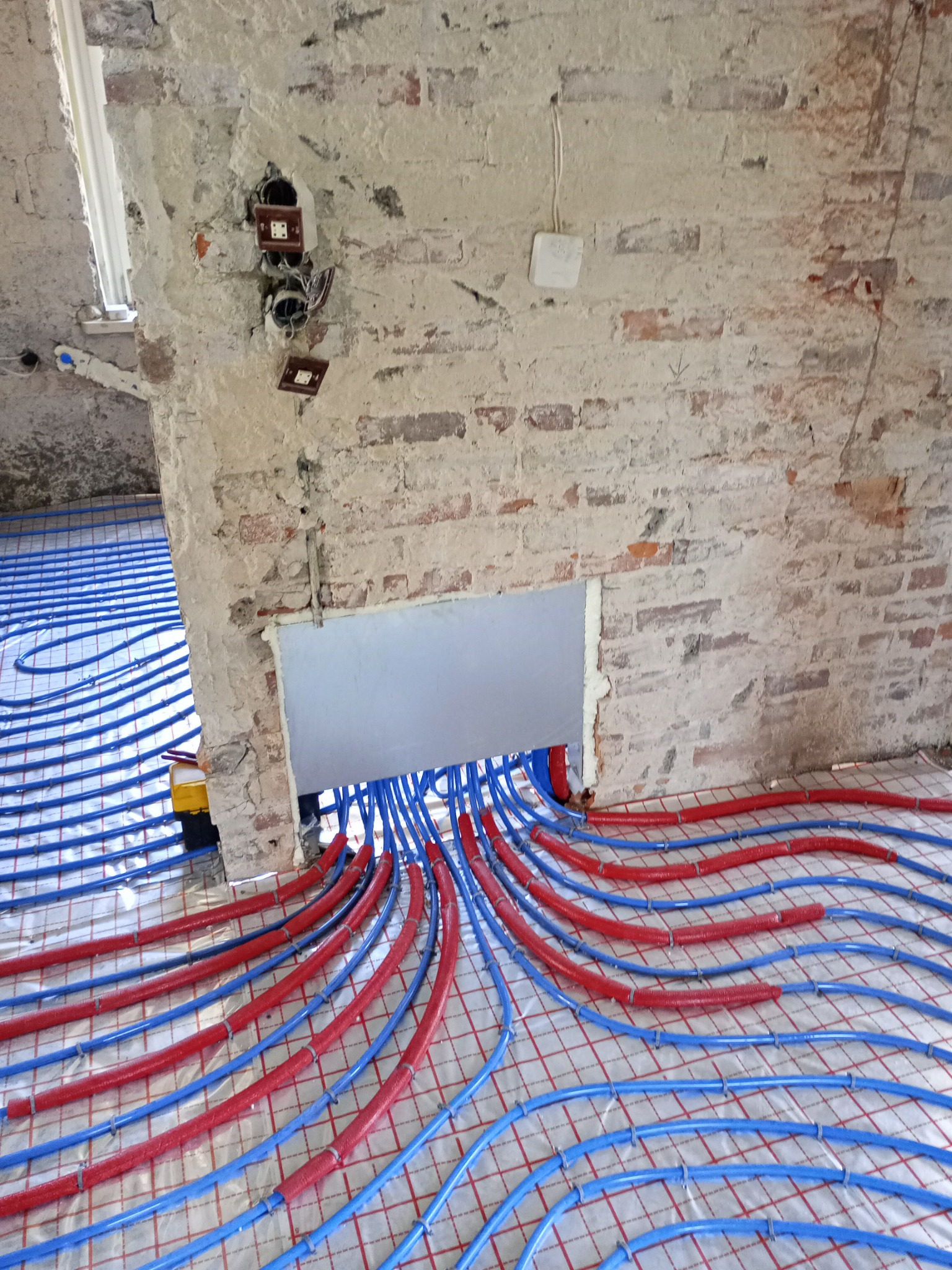 Nietypowe ujęcie instalacji ogrzewania podłogowego z rurami w kolorach niebieskim i czerwonym, w surowym wnętrzu z odsłoniętą cegłą i widoczną instalacją elektryczną.