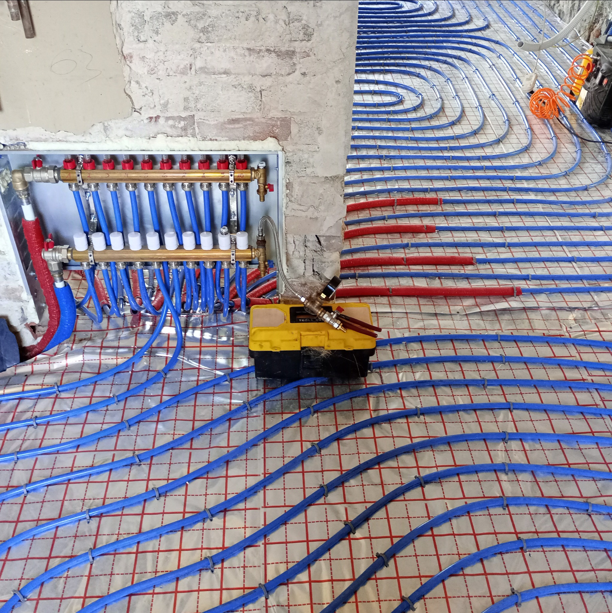 Rozłożona instalacja ogrzewania podłogowego z niebieskimi rurami na folii izolacyjnej, widoczna skrzynka rozdzielcza z czerwonymi i białymi elementami oraz żółta skrzynka narzędziowa.