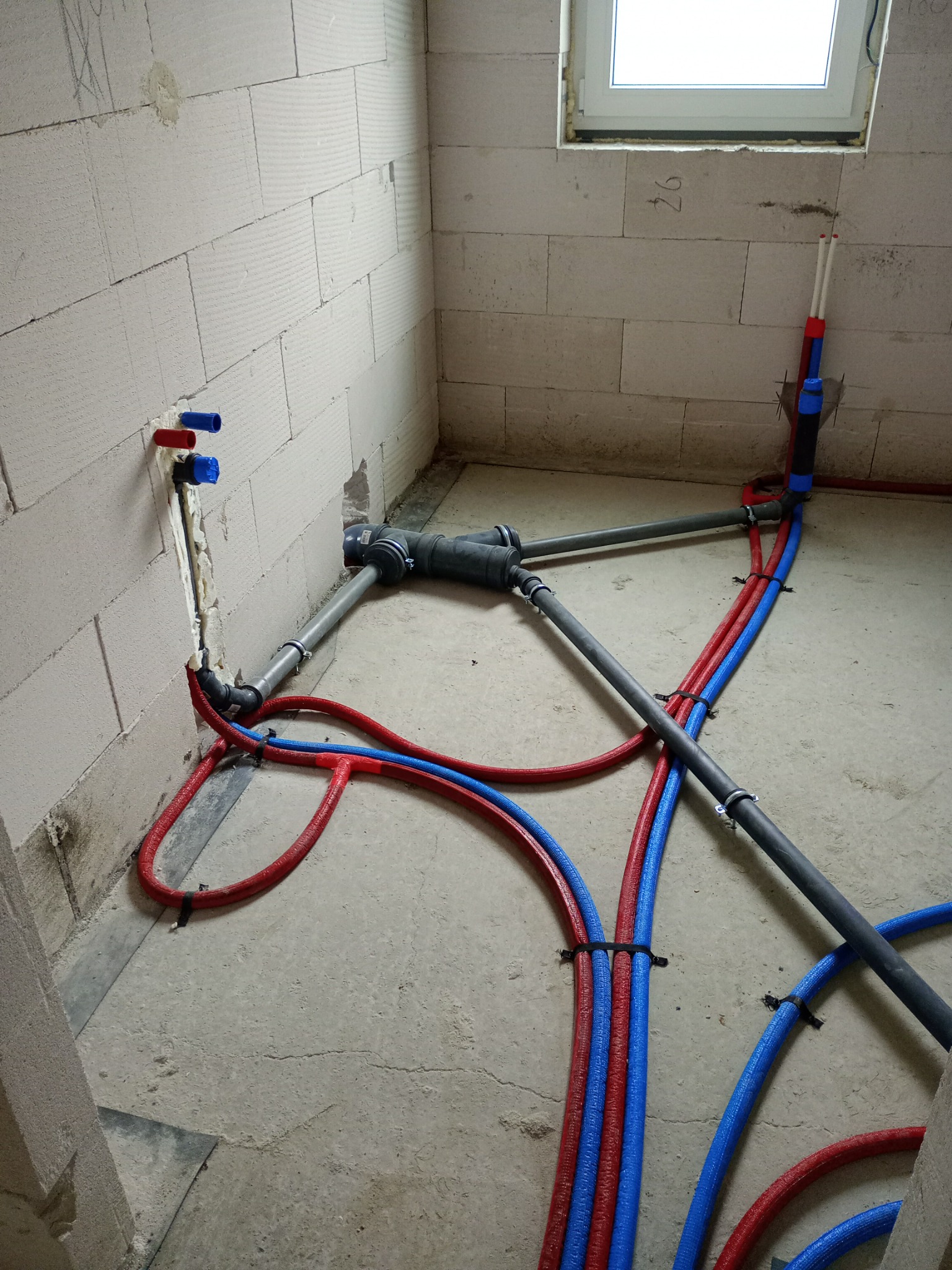 Wnętrze w trakcie montażu instalacji wodnej i kanalizacyjnej. Widoczne rury w kolorze niebieskim i czerwonym, przymocowane do ściany z betonu komórkowego.