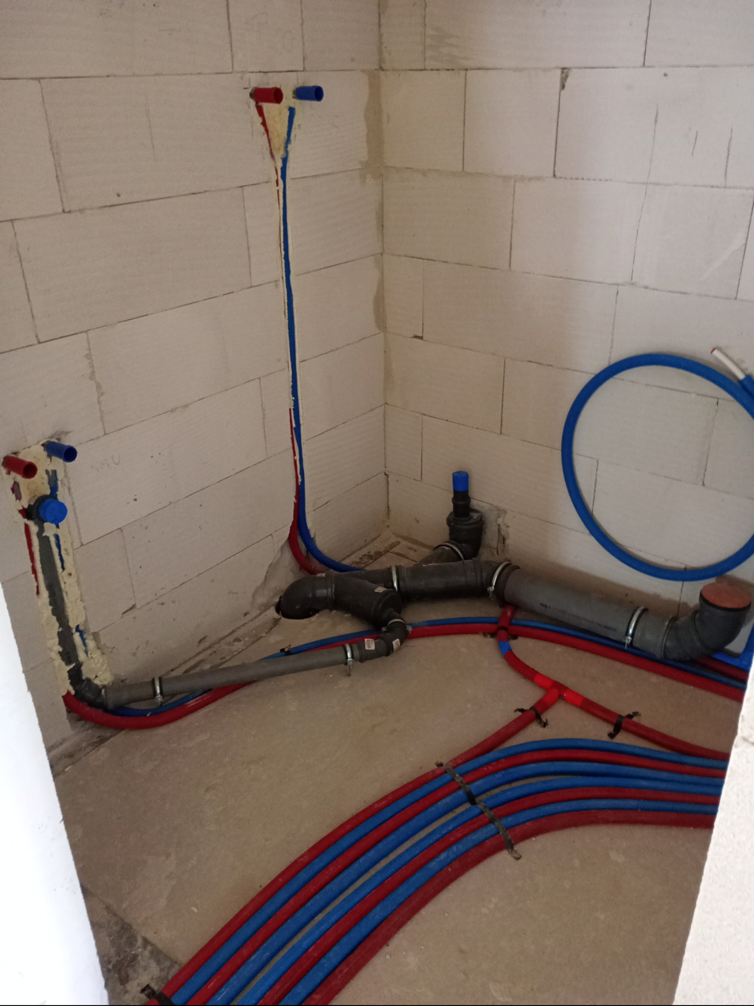Instalacja wodna i ogrzewanie podłogowe w surowym wnętrzu. Czerwone i niebieskie rury na podłodze, pionowe rury na ścianach zabezpieczone pianką montażową.