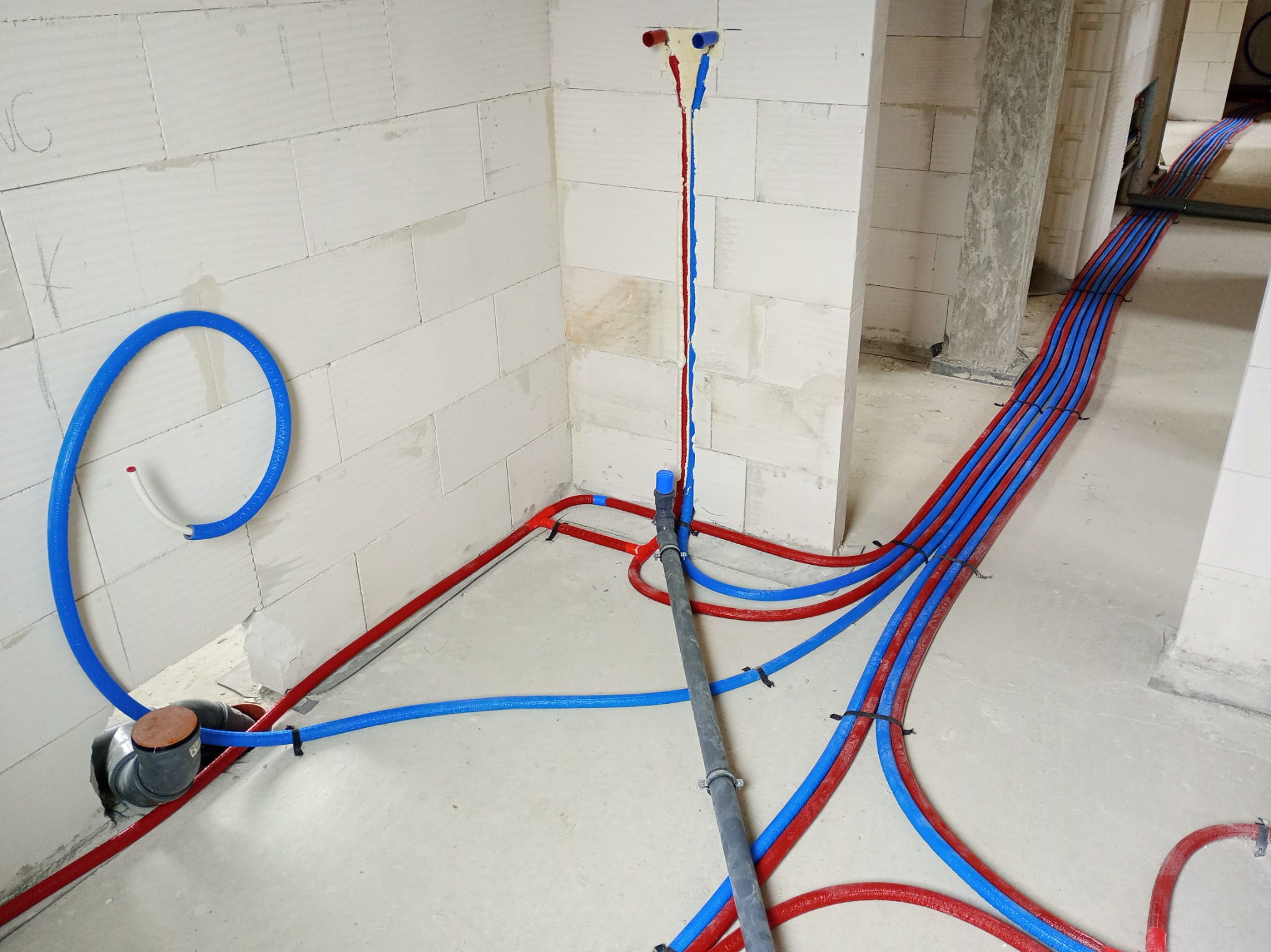 Instalacja ogrzewania podłogowego w surowym wnętrzu: rury czerwone i niebieskie prowadzone równolegle, mocowane do podłoża, zasilanie wyprowadzone na ścianie.