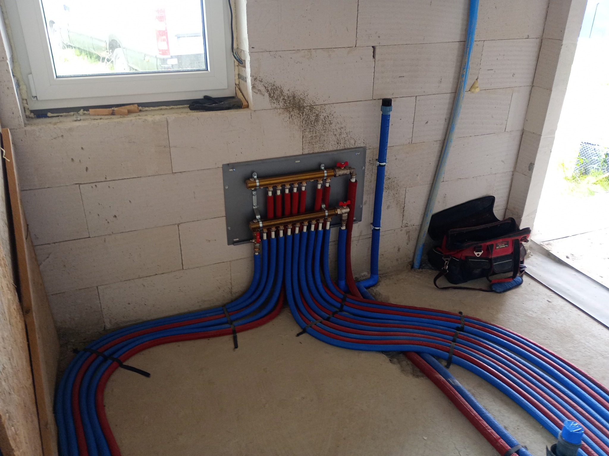 Rozdzielacz ogrzewania podłogowego z rurami w kolorze niebieskim i czerwonym, zamontowany na ścianie z bloczków betonowych w surowym wnętrzu budynku.