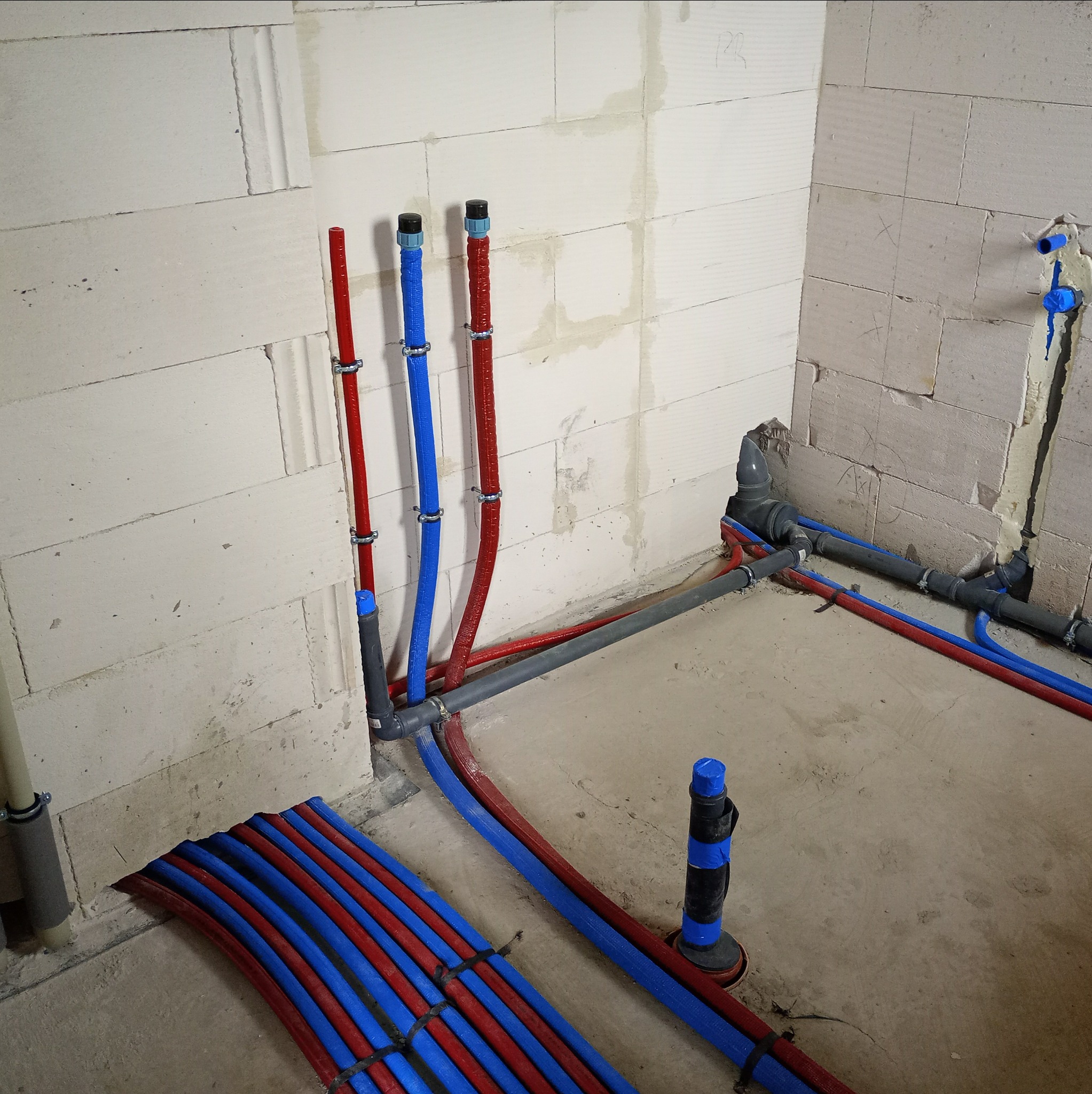 Instalacja grzewcza podłogowa w surowym wnętrzu: czerwone i niebieskie rury prowadzone wzdłuż ściany z betonu komórkowego, mocowane metalowymi uchwytami.
