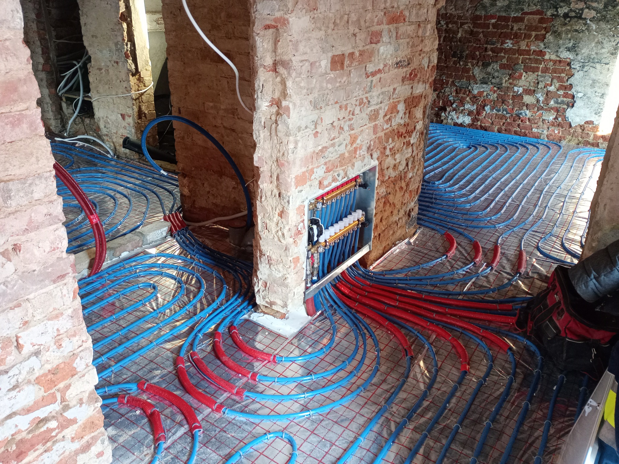 Instalacja ogrzewania podłogowego: widok na rozłożone niebieskie i czerwone rury na folii izolacyjnej, z rozdzielaczem w surowej ceglanej ścianie.