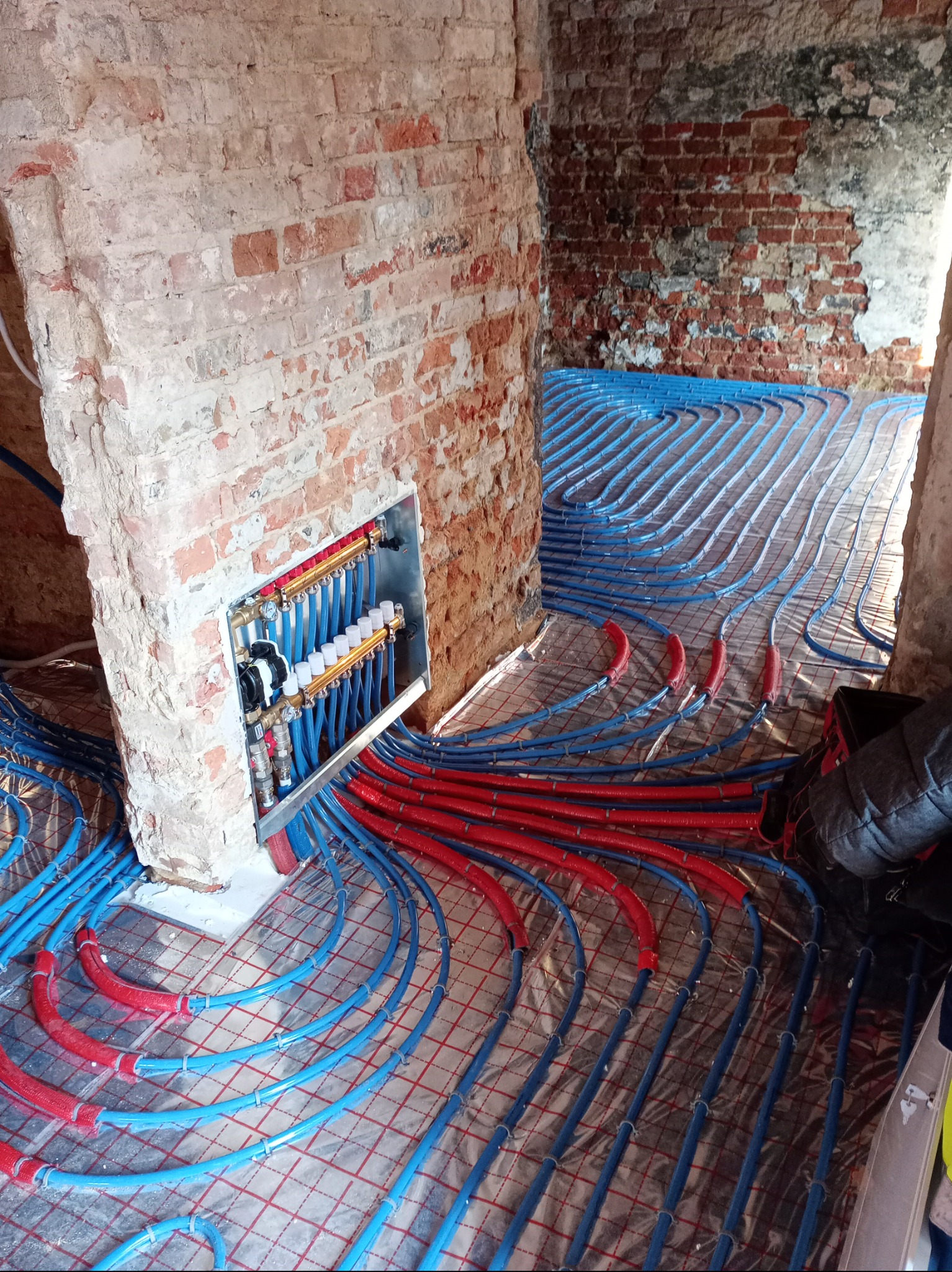 Rozłożone rury ogrzewania podłogowego (niebieskie i czerwone) na folii izolacyjnej w surowym wnętrzu z ceglanymi ścianami. Widoczna rozdzielnica w ścianie.