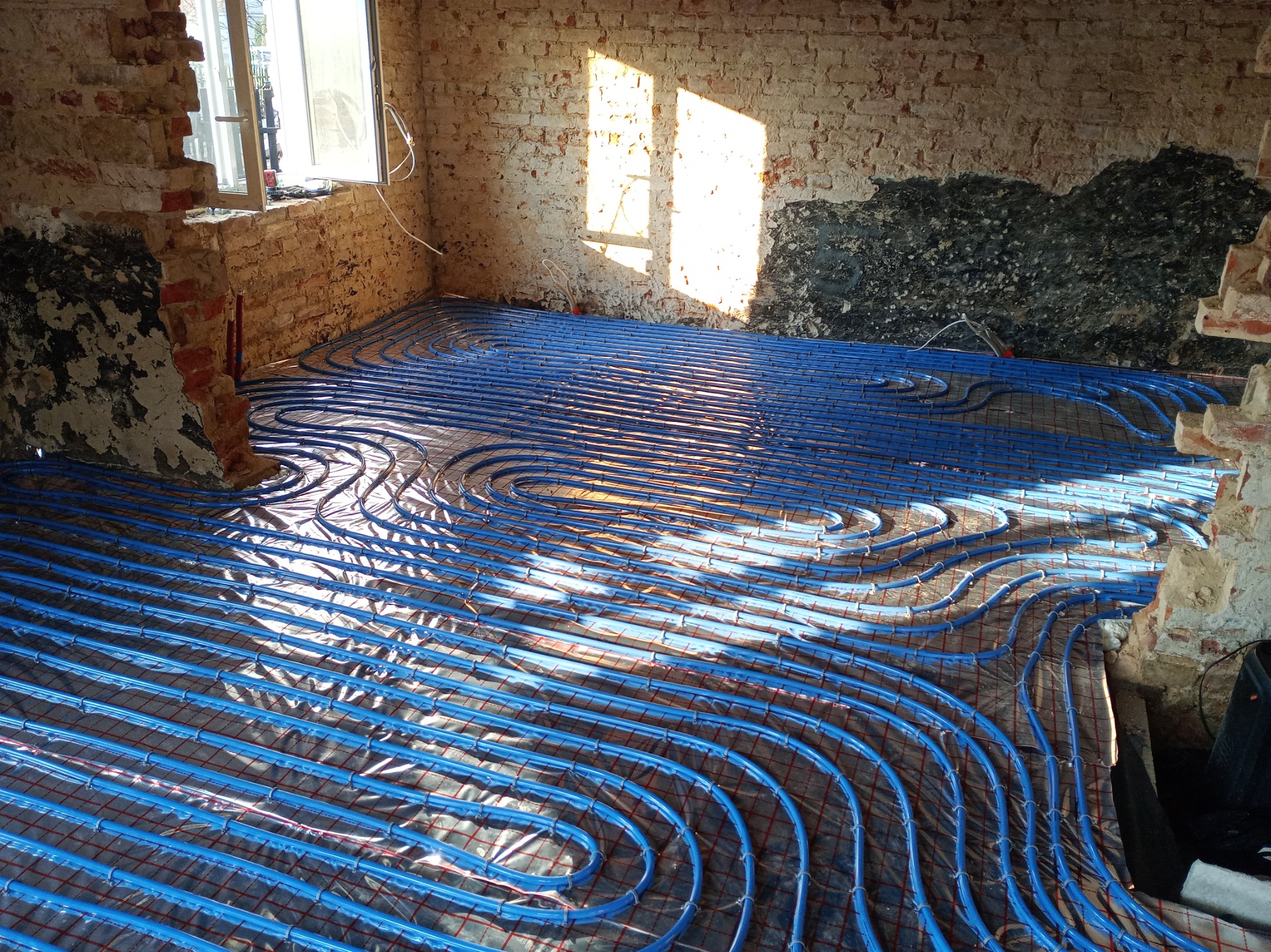 Niebieskie rury ogrzewania podłogowego ułożone na folii odblaskowej, widoczne surowe ceglane ściany w remontowanym pomieszczeniu, oświetlone promieniami słońca.