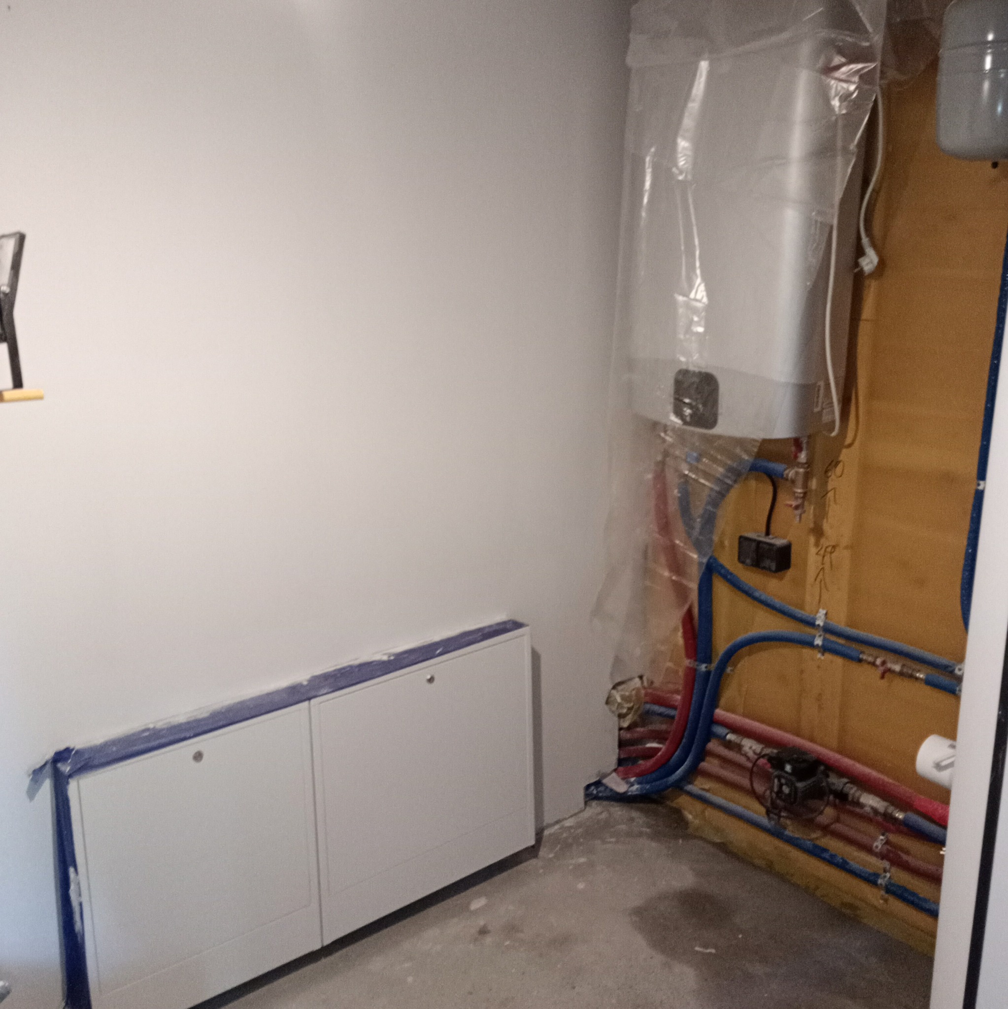 Biała szafka i bojler zabezpieczone folią ochronną podczas instalacji, widoczne rury w kolorze niebieskim i czerwonym. Ściany w kolorze białym, podłoga szara.