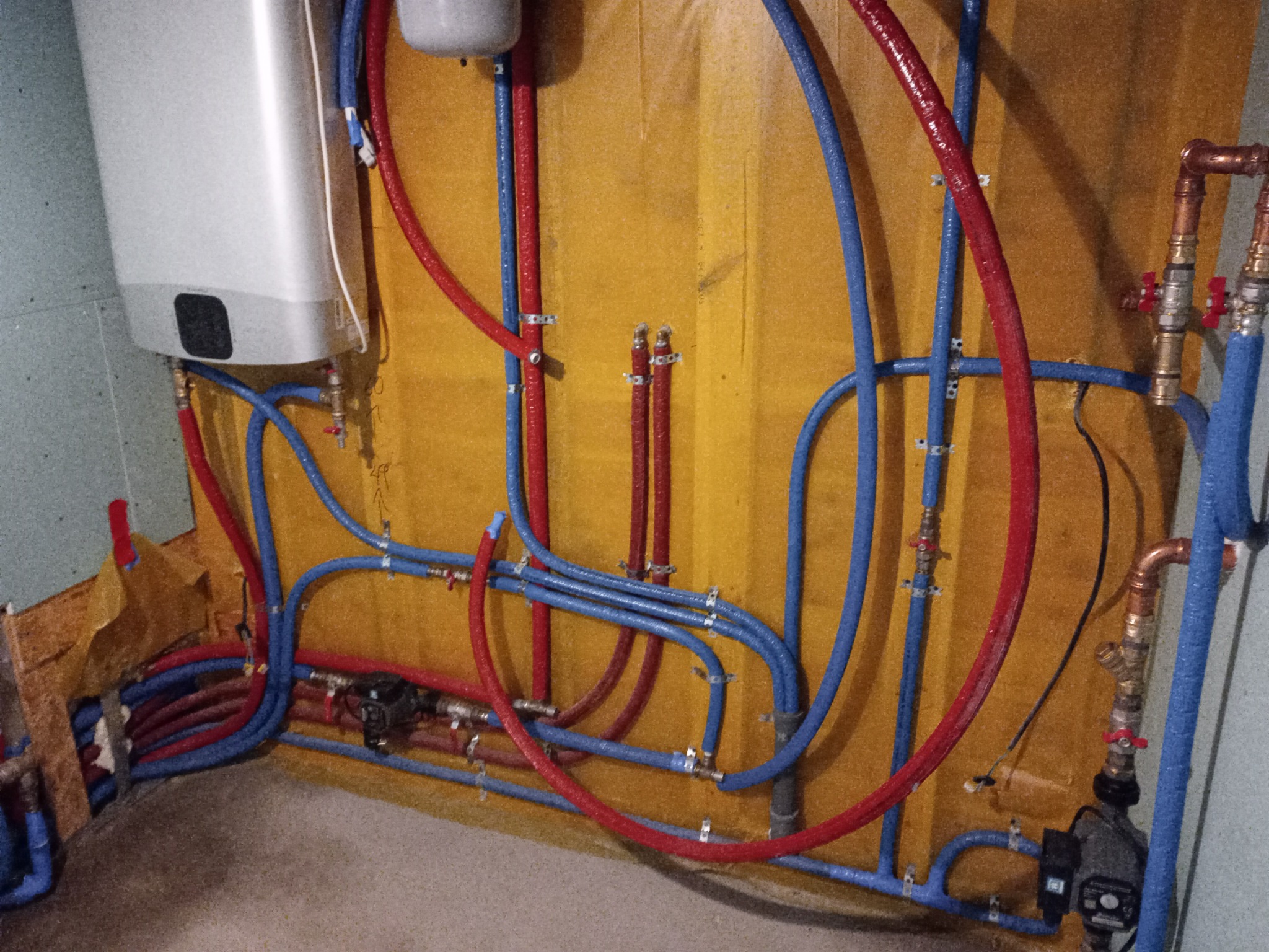 Instalacja CO i CWU z rur PEX w kolorach czerwonym i niebieskim, zamontowana na żółtej ścianie. Widoczny bojler i pompa obiegowa. Elementy instalacji hydraulicznej.