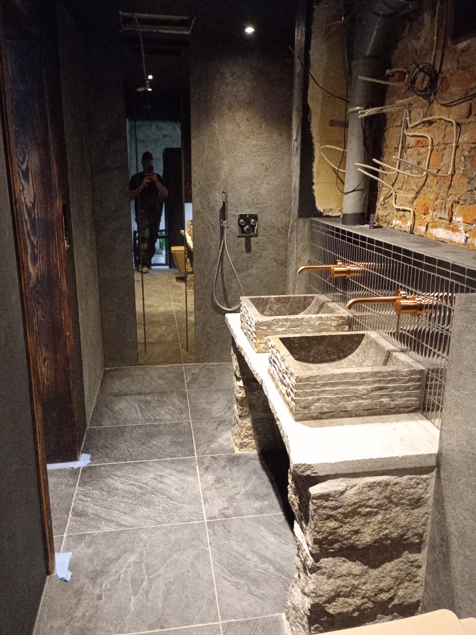 Nowoczesna łazienka z dwoma kamiennymi umywalkami i miedzianymi kranami. W tle lustro z odbiciem osoby robiącej zdjęcie. Widoczna instalacja na ścianie z cegły.