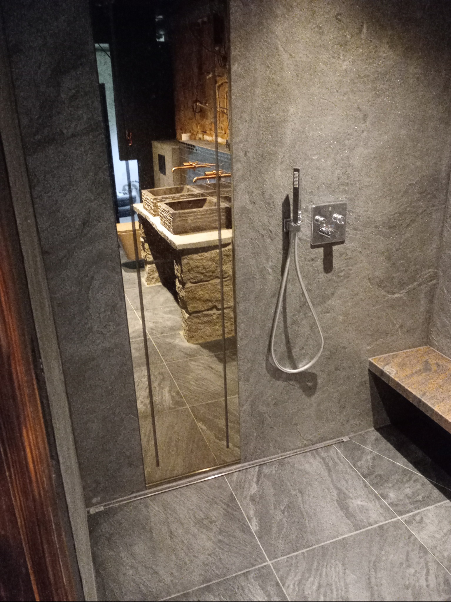 Nowoczesna łazienka z kamienną ścianą i kabiną prysznicową. Widoczny panel sterujący prysznicem i kamienna ławeczka. W lustrze odbija się umywalka z kamienia.