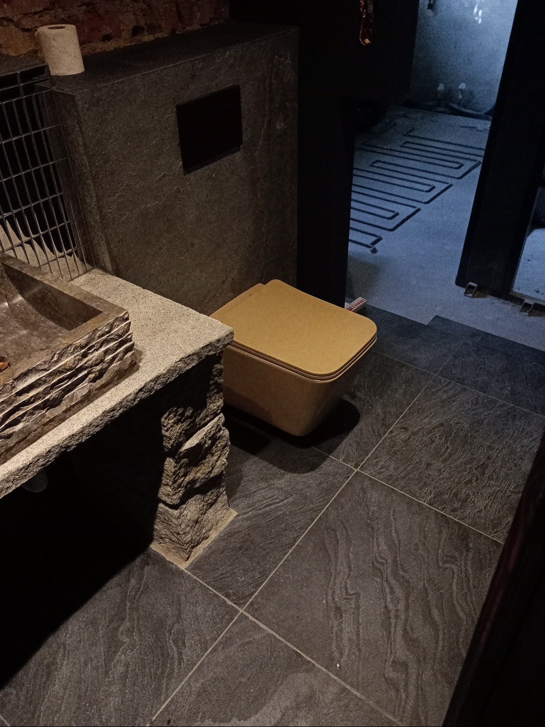 Nowoczesna łazienka z kamienną umywalką i toaletą w odcieniach beżu i szarości. W tle widoczna instalacja ogrzewania podłogowego.