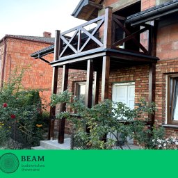 BEAM Michał Łoniewski - Drewniana zabudowa balkonu z balustradą krzyżową, ciemne drewno na tle ceglanej elewacji, widok z dołu, częściowo zasłonięta roślinnością.