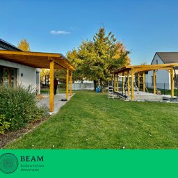 BEAM Michał Łoniewski - Dwie drewniane pergole w trakcie budowy na zielonym trawniku, z elementami konstrukcyjnymi i drabinką. W tle domy i drzewa pod błękitnym niebem.