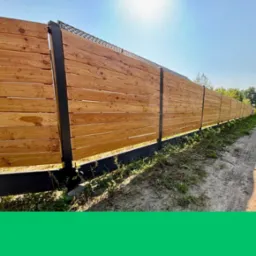 Długi, drewniany płot z poziomych desek i czarnych słupków, oddzielający posesję od drogi. Widoczne ślady opon na ziemi i zieleń. Słoneczny dzień.