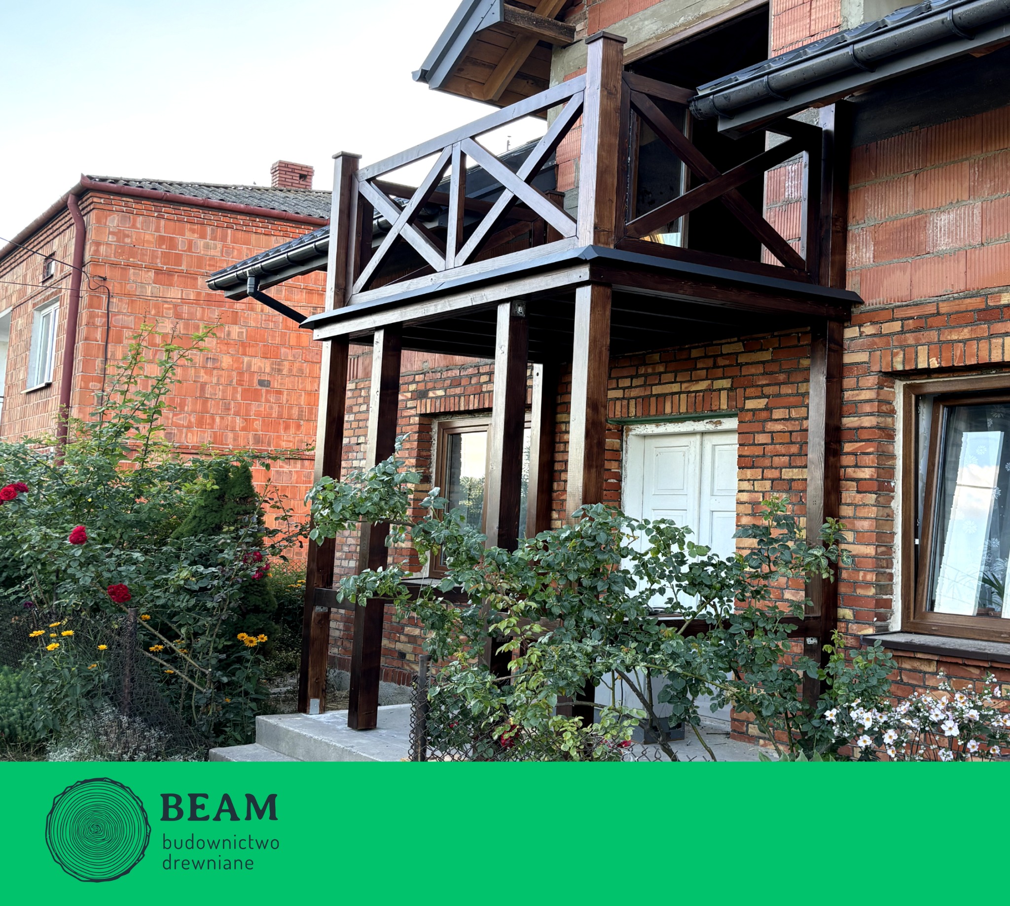 Drewniana zabudowa balkonu z balustradą krzyżową, ciemne drewno na tle ceglanej elewacji, widok z dołu, częściowo zasłonięta roślinnością.
