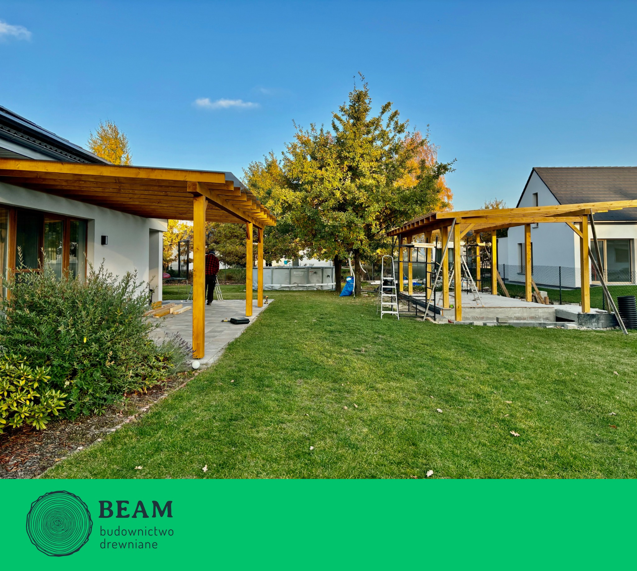 Dwie drewniane pergole w trakcie budowy na zielonym trawniku, z elementami konstrukcyjnymi i drabinką. W tle domy i drzewa pod błękitnym niebem.