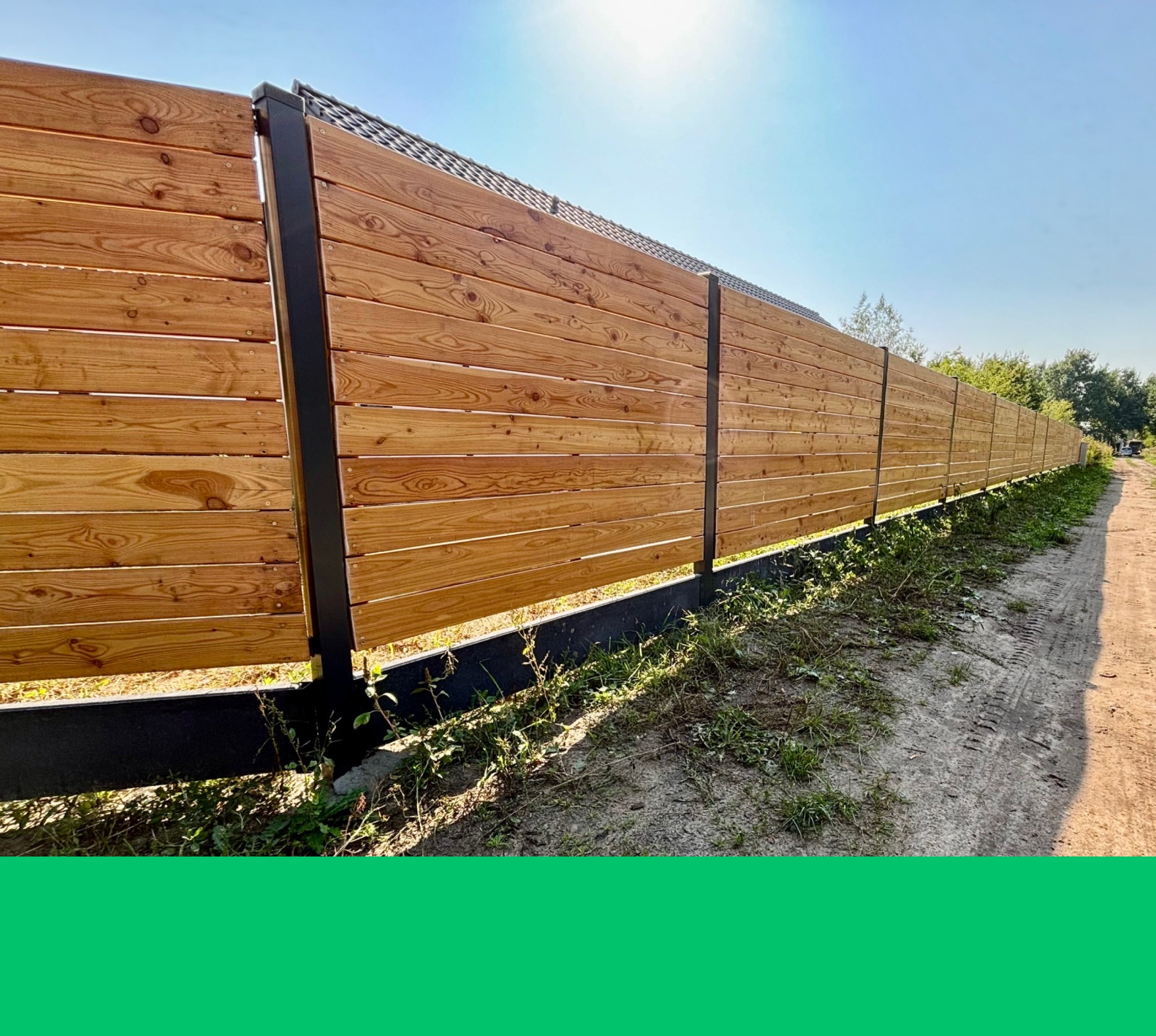 Długi, drewniany płot z poziomych desek i czarnych słupków, oddzielający posesję od drogi. Widoczne ślady opon na ziemi i zieleń. Słoneczny dzień.