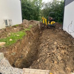 Aftergarden- Ogrody Premium - Żółta mini koparka w wykopie między dwoma białymi budynkami, operator pracuje nad wyrównywaniem terenu pod przyszły ogród lub instalację.