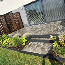 Aftergarden- Ogrody Premium - Nowoczesny ogród z drewnianą ażurową przesłoną, schodami z kostki brukowej i rabatami z różnorodną roślinnością przy budynku.