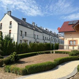 Aftergarden- Ogrody Premium - Przygotowany teren pod trawnik otoczony niskim żywopłotem, na tle szeregowej zabudowy i domu z czerwoną dachówką oraz ozdobnie przyciętymi iglakami.