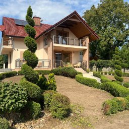Aftergarden- Ogrody Premium - Uformowany w spiralę iglak przed nowoczesnym domem z czerwonym dachem, otoczony niskim żywopłotem i jasną ścieżką z kostki brukowej, częściowo przygotowany teren pod nasadzenia.