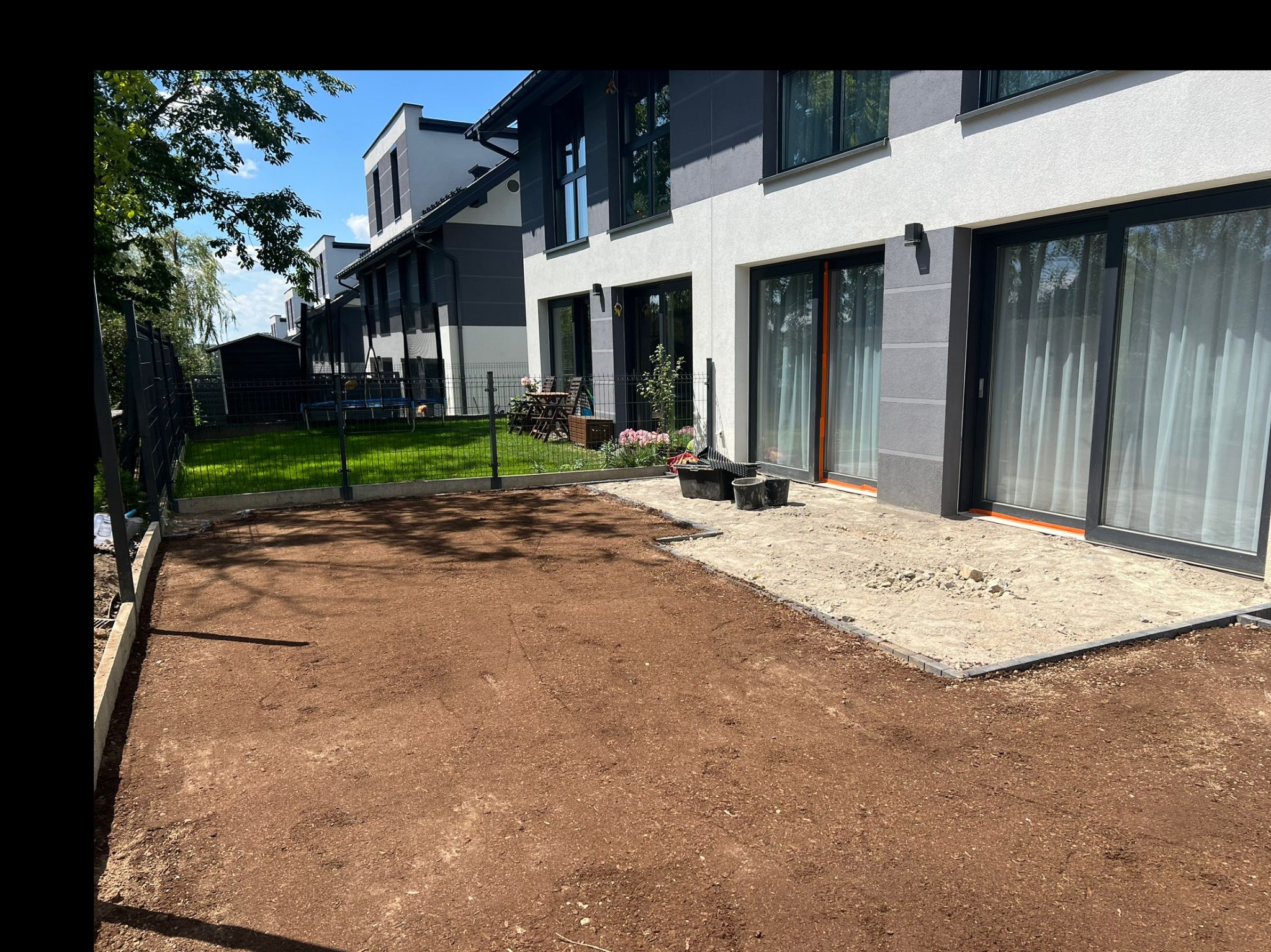 Przygotowany teren pod trawnik z wyrównaną ziemią i obrzeżami, przylegający do tarasu z piaskiem, nowoczesny dom w tle.