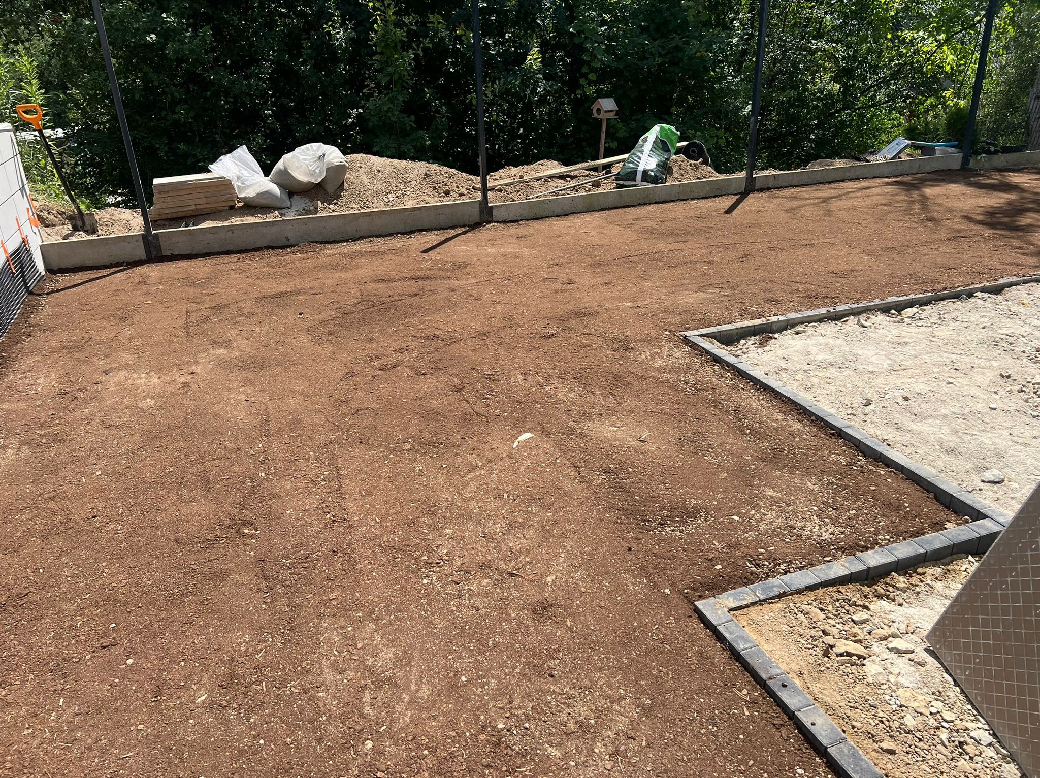 Przygotowany teren pod przyszły trawnik z rozłożoną ziemią, obrzeżami i widocznymi narzędziami ogrodniczymi w tle.