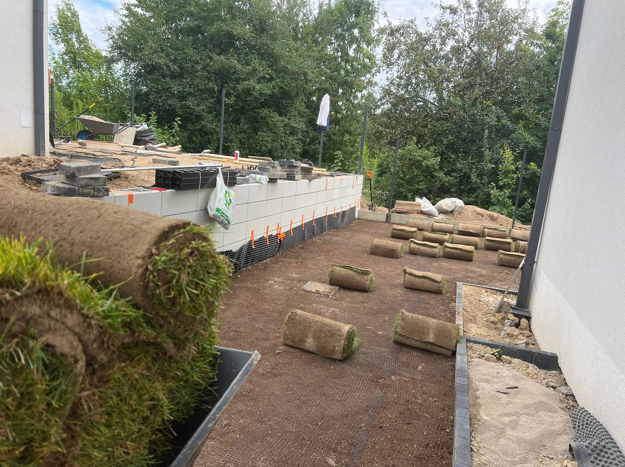 Rolki trawy w taczce i rozłożone na przygotowanym gruncie, widoczna podbudowa muru oporowego i narzędzia w tle, teren w trakcie prac ziemnych.