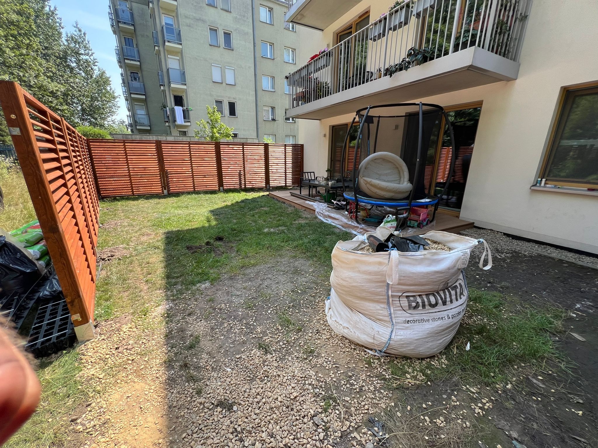 Teren w trakcie zagospodarowywania: widoczny fragment nowego drewnianego płotu lamelowego, wysypany żwir, duży worek z kamieniami dekoracyjnymi i fragment trawnika przed budynkiem wielorodzinnym.