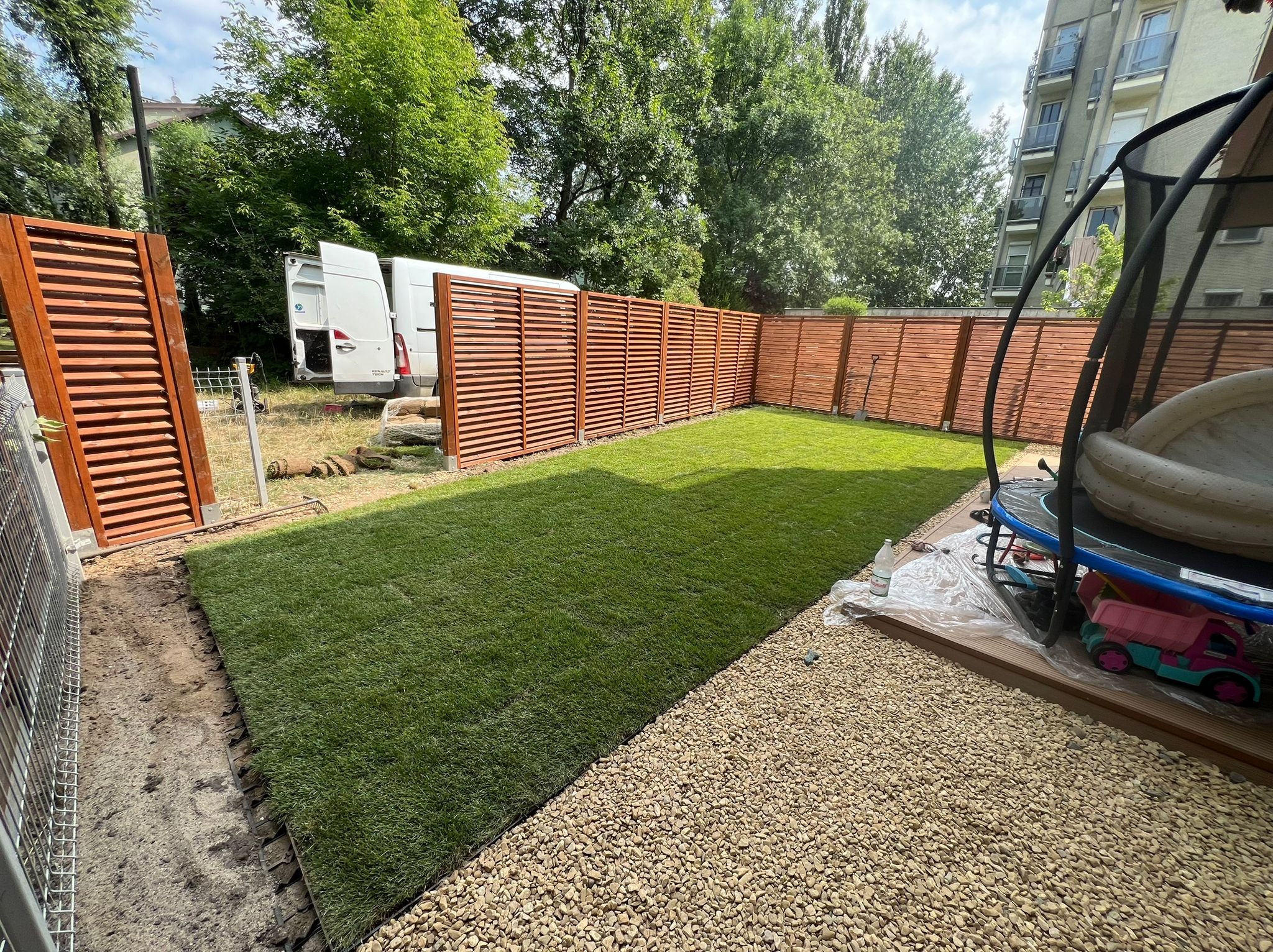 Świeżo ułożona, soczysta darń w ogrodzie z drewnianym ogrodzeniem lamelowym; w tle biały van z otwartymi drzwiami i sprzęt ogrodowy.