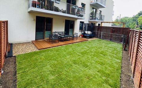 Świeżo założony trawnik z rolki przylegający do tarasu z desek kompozytowych w ogrodzie miejskim, widoczny fragment brązowego płotu lamelowego i trampolina w tle.