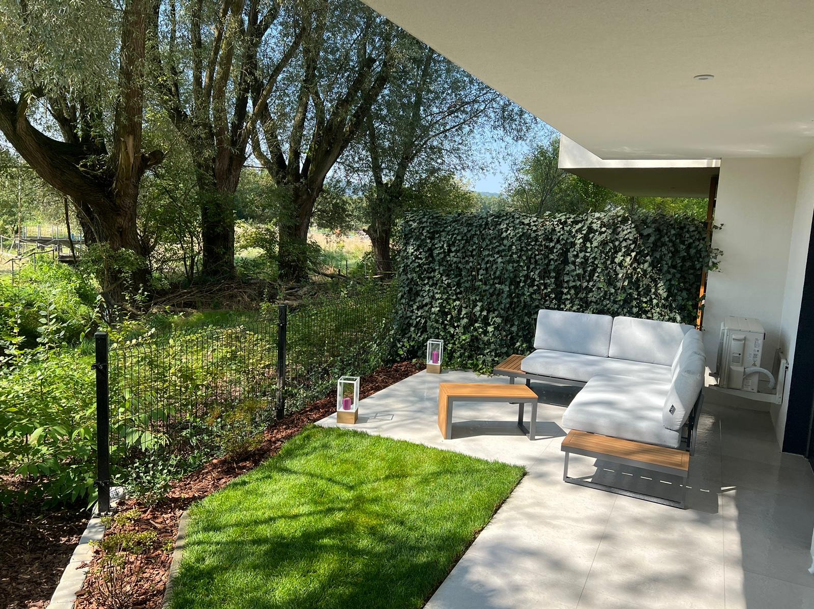 Nowoczesny taras z narożną sofą z jasnymi poduszkami, drewnianym stolikiem kawowym i fragmentem trawnika z rolki, otoczony siatką ogrodzeniową i żywopłotem z bluszczu.