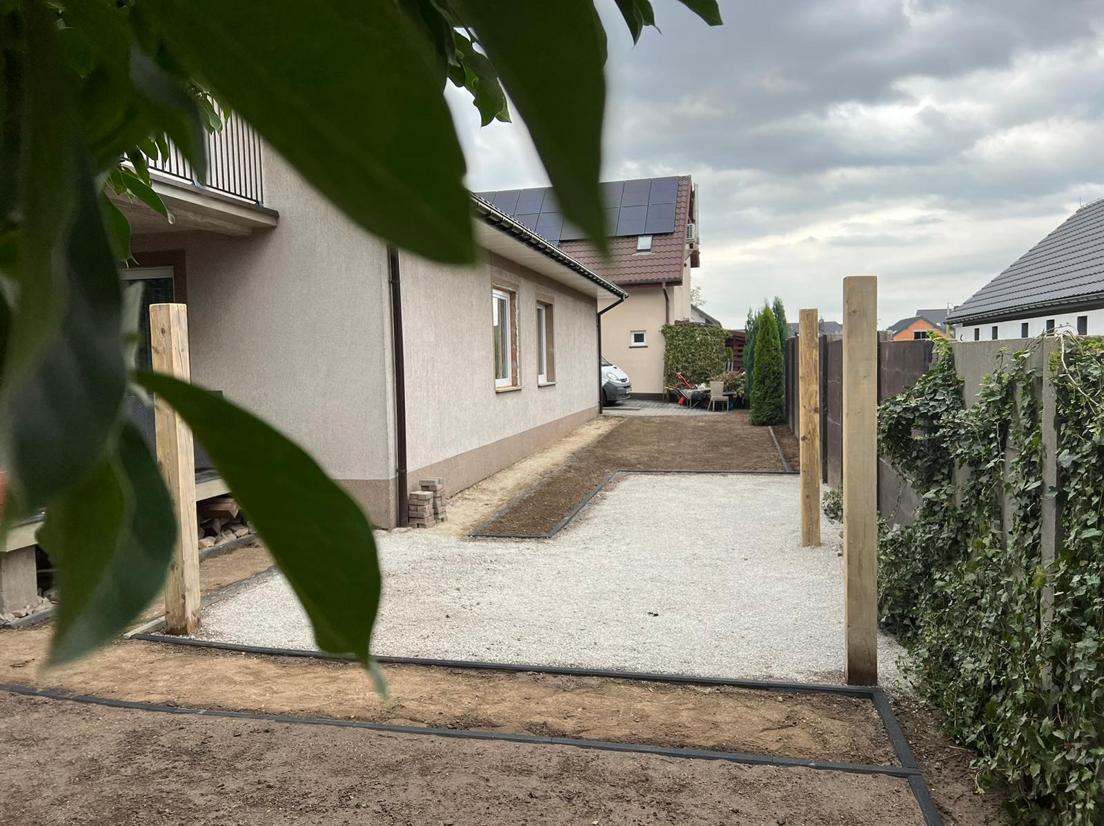 Przygotowany teren pod ogród: wysypany żwir, obrzeża z tworzywa, widoczne słupki ogrodzenia, trawnik w tle, fragment domu i roślinność w kadrze.