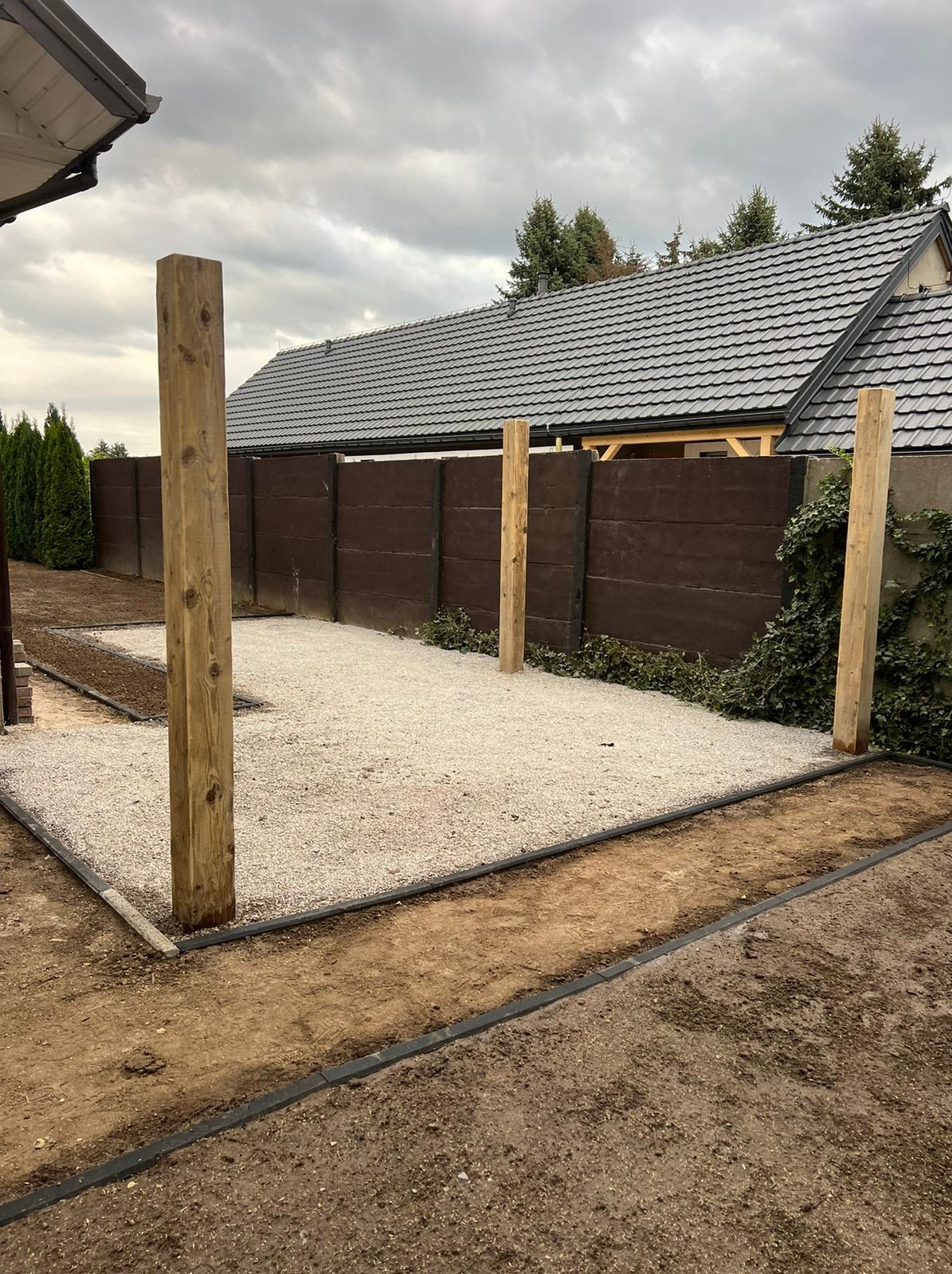 Przygotowany teren pod budowę altany lub pergoli: wysypany żwir, ustawione drewniane słupy nośne, obrzeża trawnika z tworzywa.