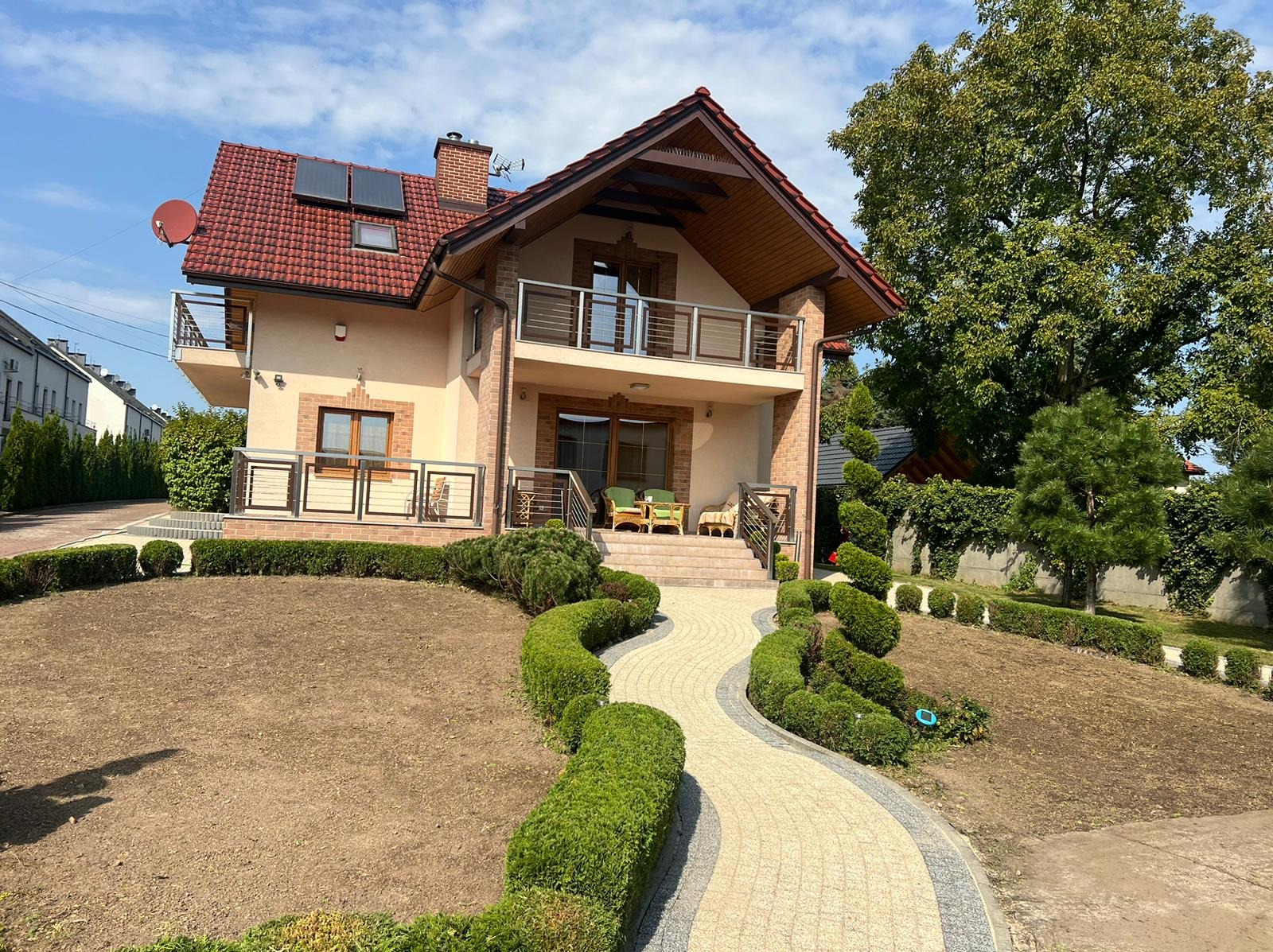 Dwupiętrowy dom z beżową elewacją, czerwoną dachówką i balkonami, otoczony ozdobnymi krzewami i ścieżką z kostki brukowej, z przygotowanym terenem pod przyszły trawnik.