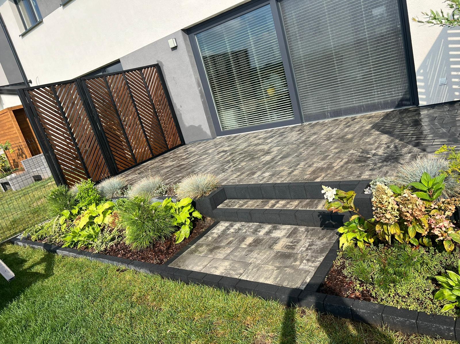Nowoczesny ogród z drewnianą ażurową przesłoną, schodami z kostki brukowej i rabatami z różnorodną roślinnością przy budynku.