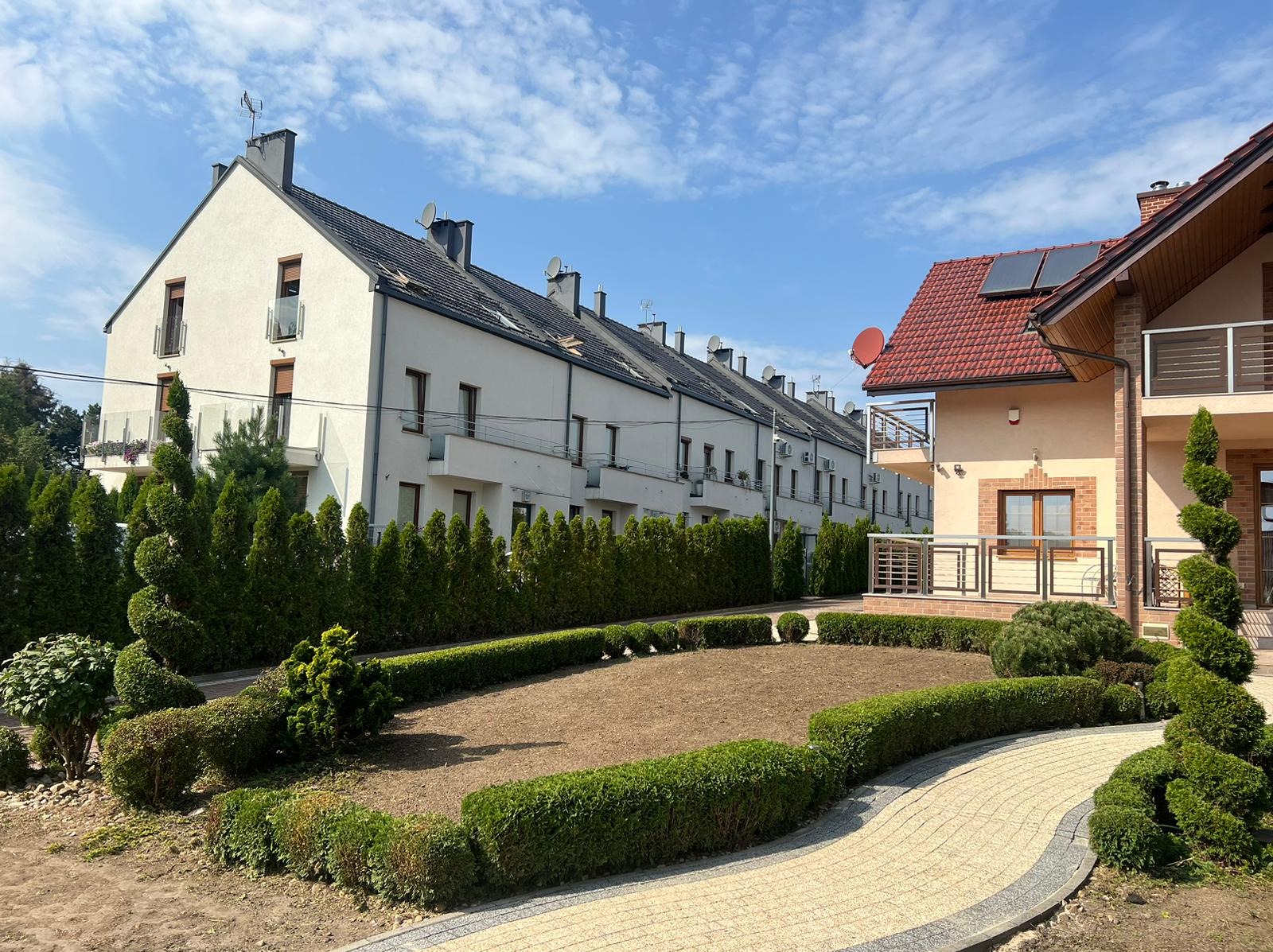 Przygotowany teren pod trawnik otoczony niskim żywopłotem, na tle szeregowej zabudowy i domu z czerwoną dachówką oraz ozdobnie przyciętymi iglakami.