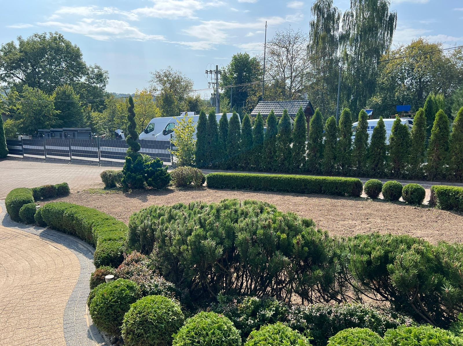 Starannie utrzymany ogród z równo przyciętymi krzewami i rzędami iglaków, widoczny trawnik z podsypanym żwirem, w tle brama i budynek.