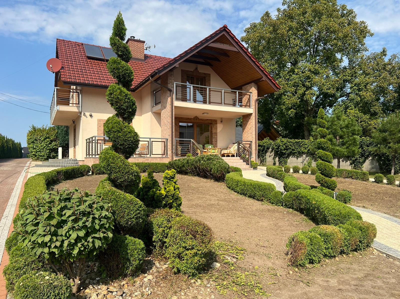 Uformowany w spiralę iglak przed nowoczesnym domem z czerwonym dachem, otoczony niskim żywopłotem i jasną ścieżką z kostki brukowej, częściowo przygotowany teren pod nasadzenia.