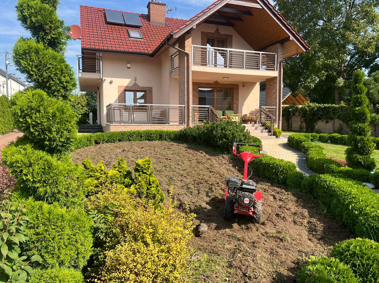 Czerwony glebogryzarek spalinowy na przygotowanym podłożu z widocznymi śladami po pracy, w tle duży dom z balkonami i zadbany ogród z tujami.