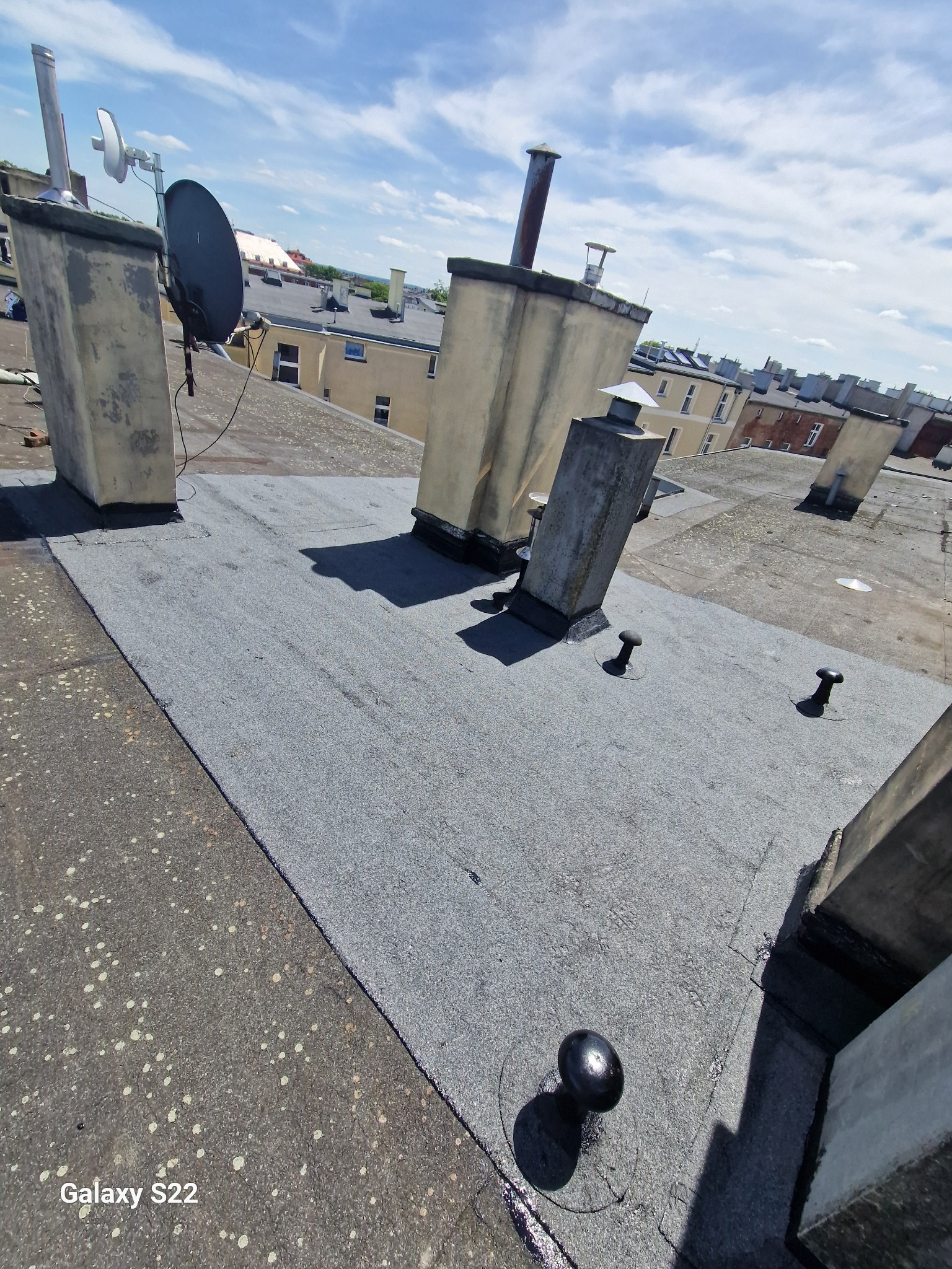 Widok z góry na płaski dach pokryty papą termozgrzewalną, z kominami i anteną satelitarną na tle zabudowy miejskiej i błękitnego nieba.