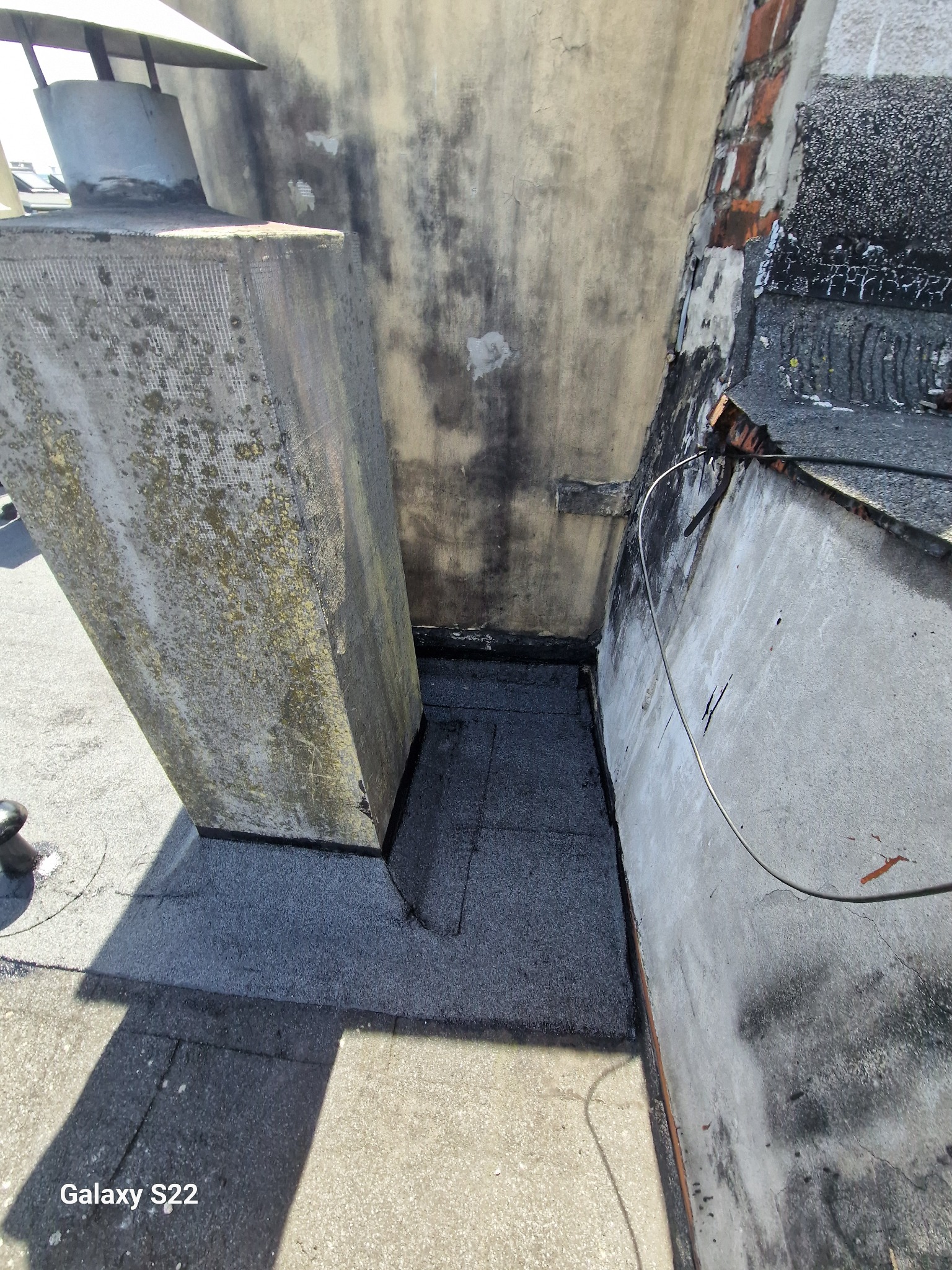 Szczegółowe ujęcie uszczelnienia dachu wokół komina z użyciem papy bitumicznej, widoczne ślady zabrudzeń i wilgoci na kominie oraz ścianie.