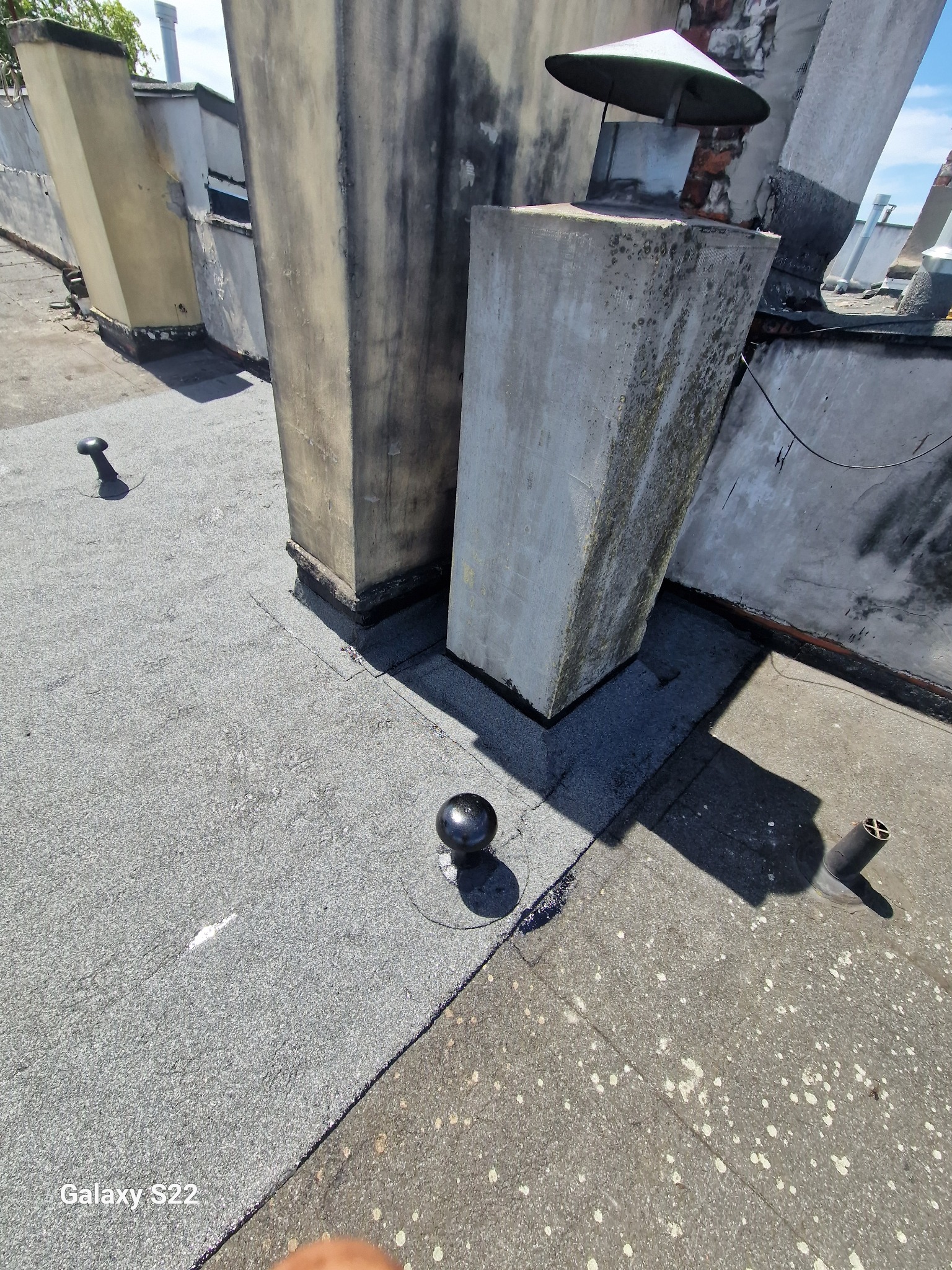 Widok z góry na fragment dachu pokrytego papą, z kominami zabezpieczonymi obróbkami blacharskimi i odpowietrznikami.