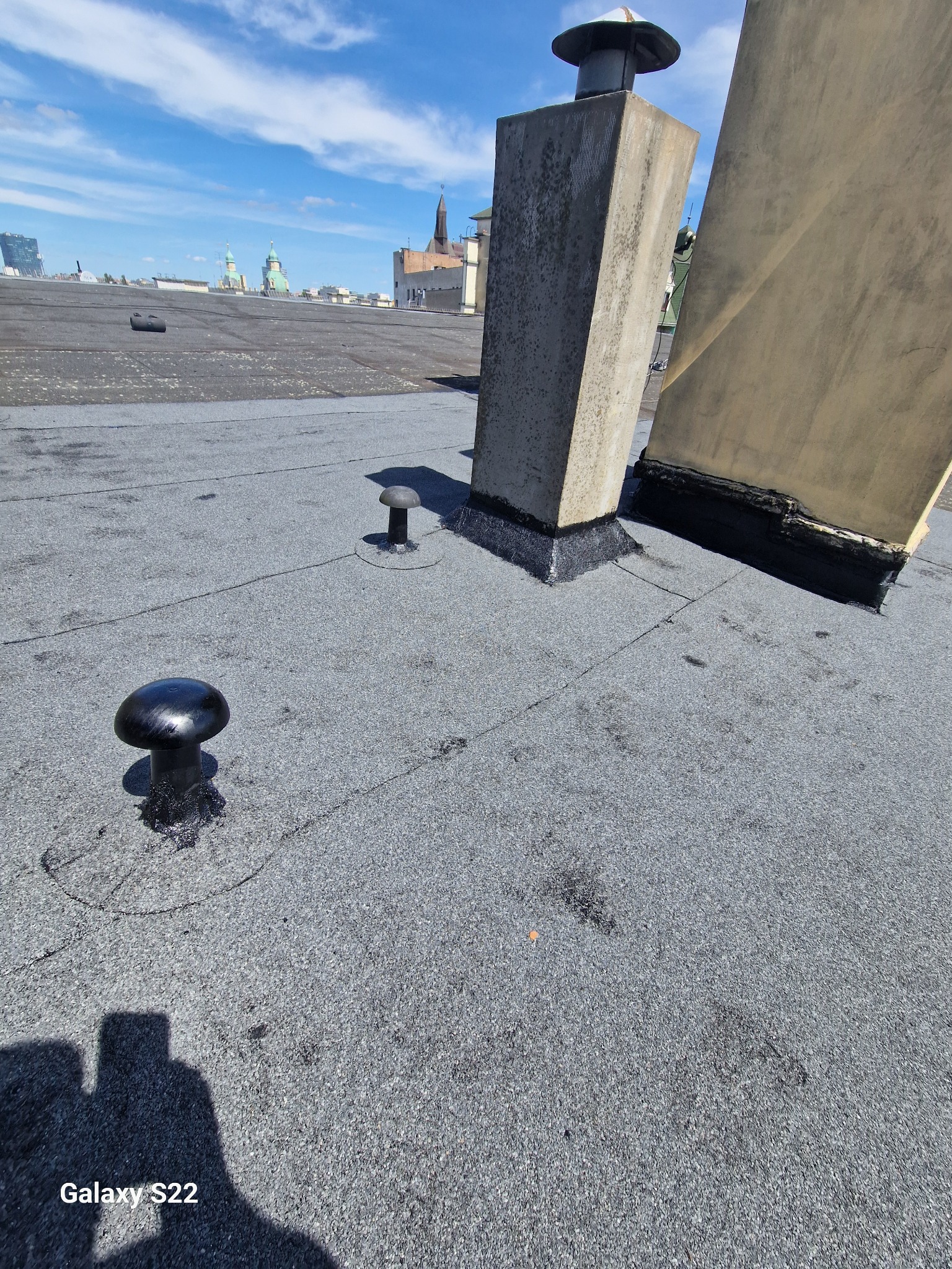 Płaski dach pokryty papą z widocznymi kominami i odpowietrznikami; w tle panorama miasta z wieżami kościołów.