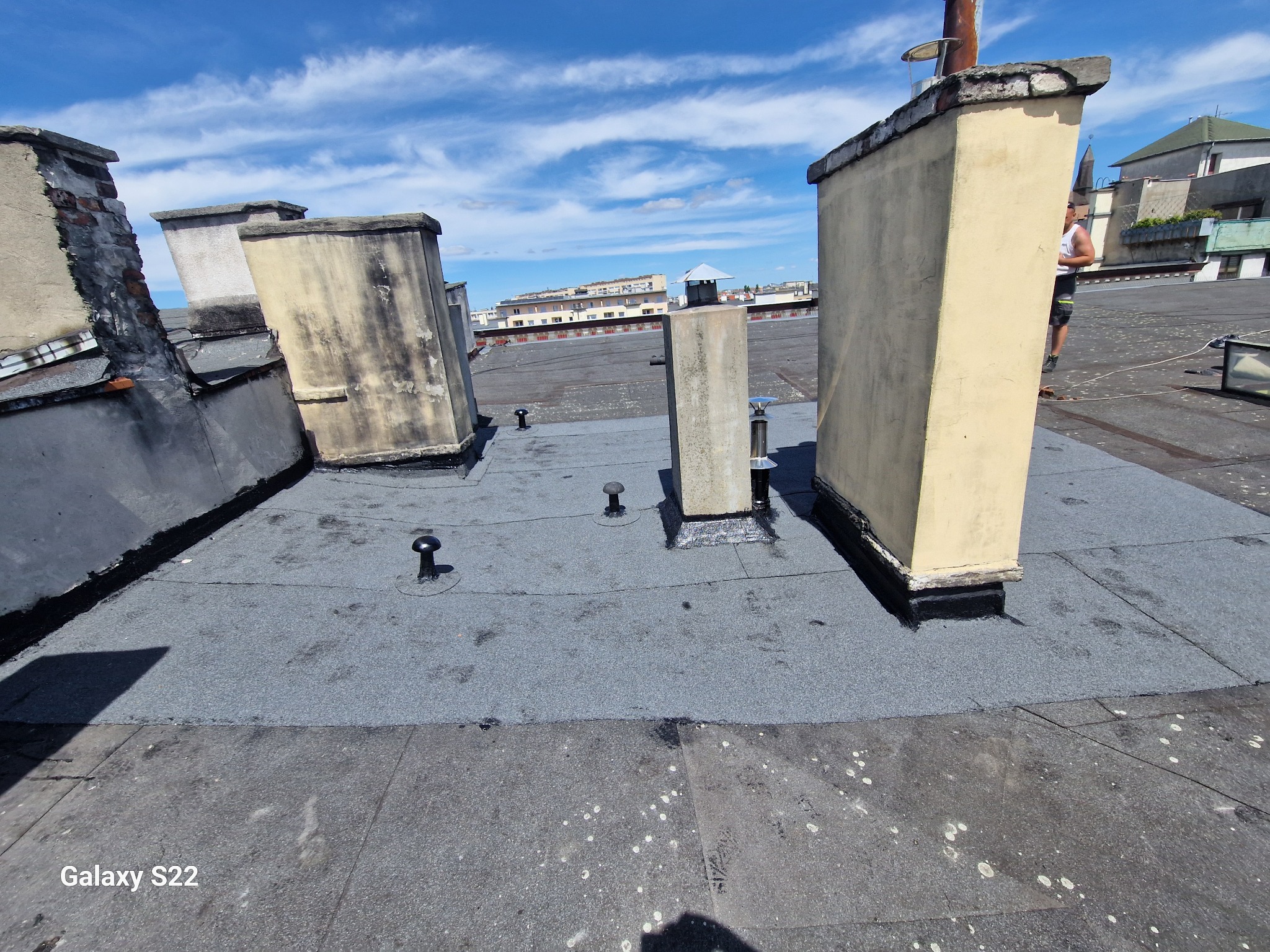 Widok z góry na płaski dach pokryty papą, z widocznymi kominami o zróżnicowanej kondycji, zarys sylwetki osoby w tle, błękitne niebo z chmurami.