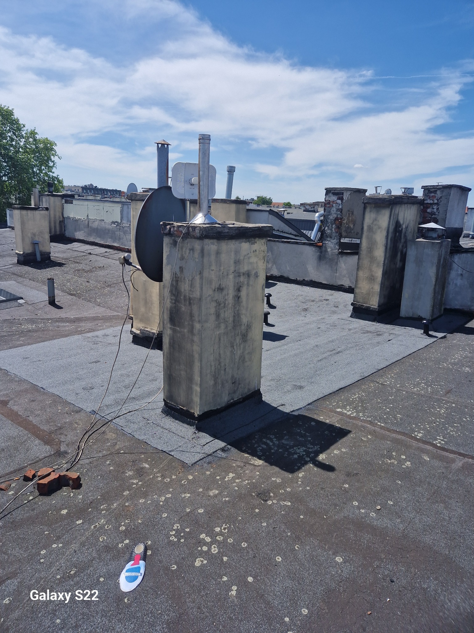 Płaski dach pokryty papą z licznymi kominami i anteną satelitarną; widoczne ślady po ptasich odchodach i pozostawione cegły.